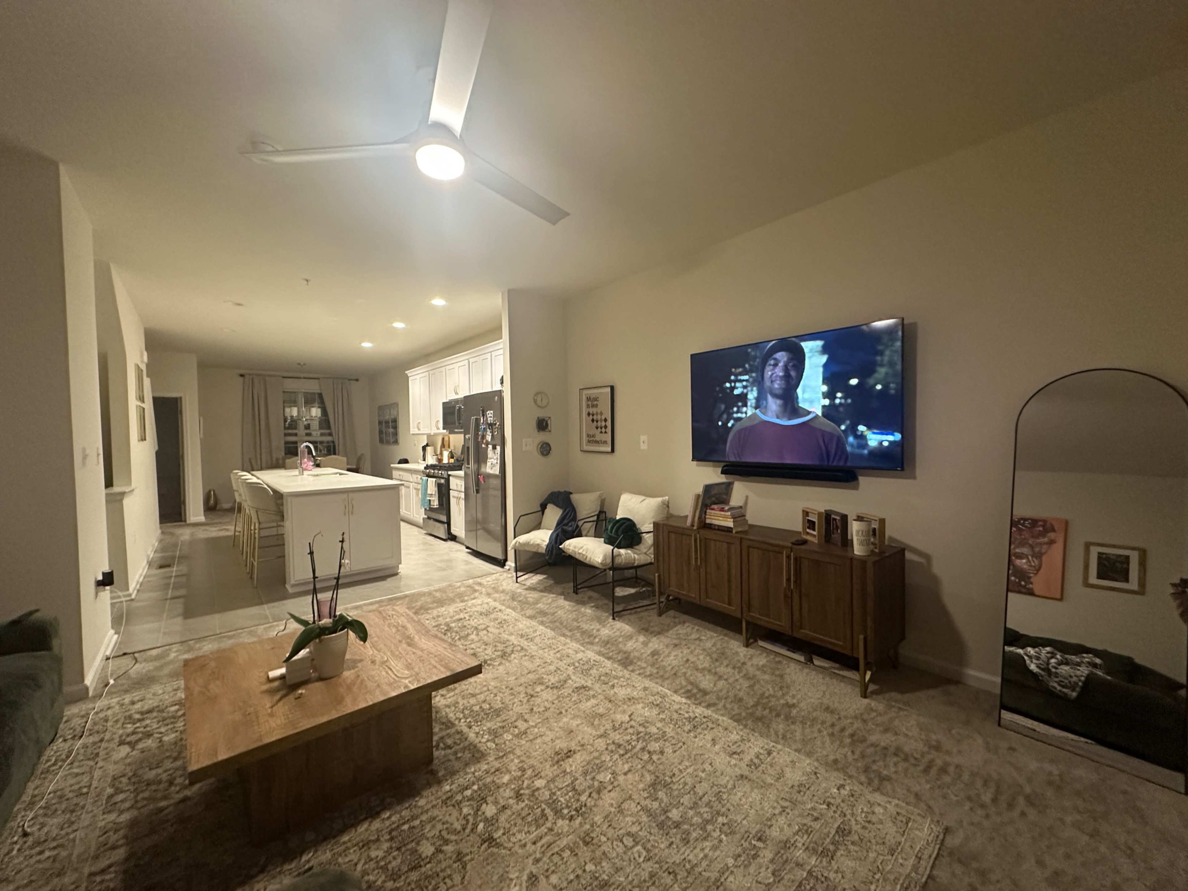 The image shows a modern living room featuring a large TV mounted on the wall, a wooden coffee table, and a view into an adjacent kitchen with white cabinets.
