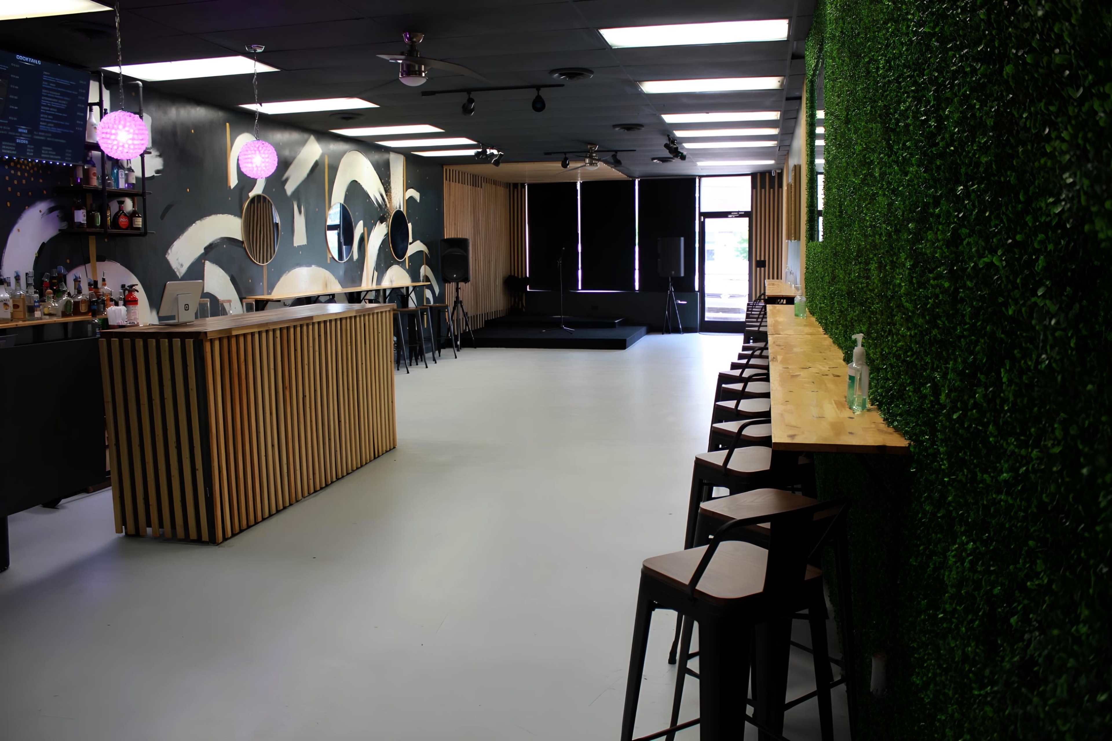 A spacious, modern interior of a bar with wooden accents, a long countertop, and green foliage on one wall, along with a small stage area at the back.