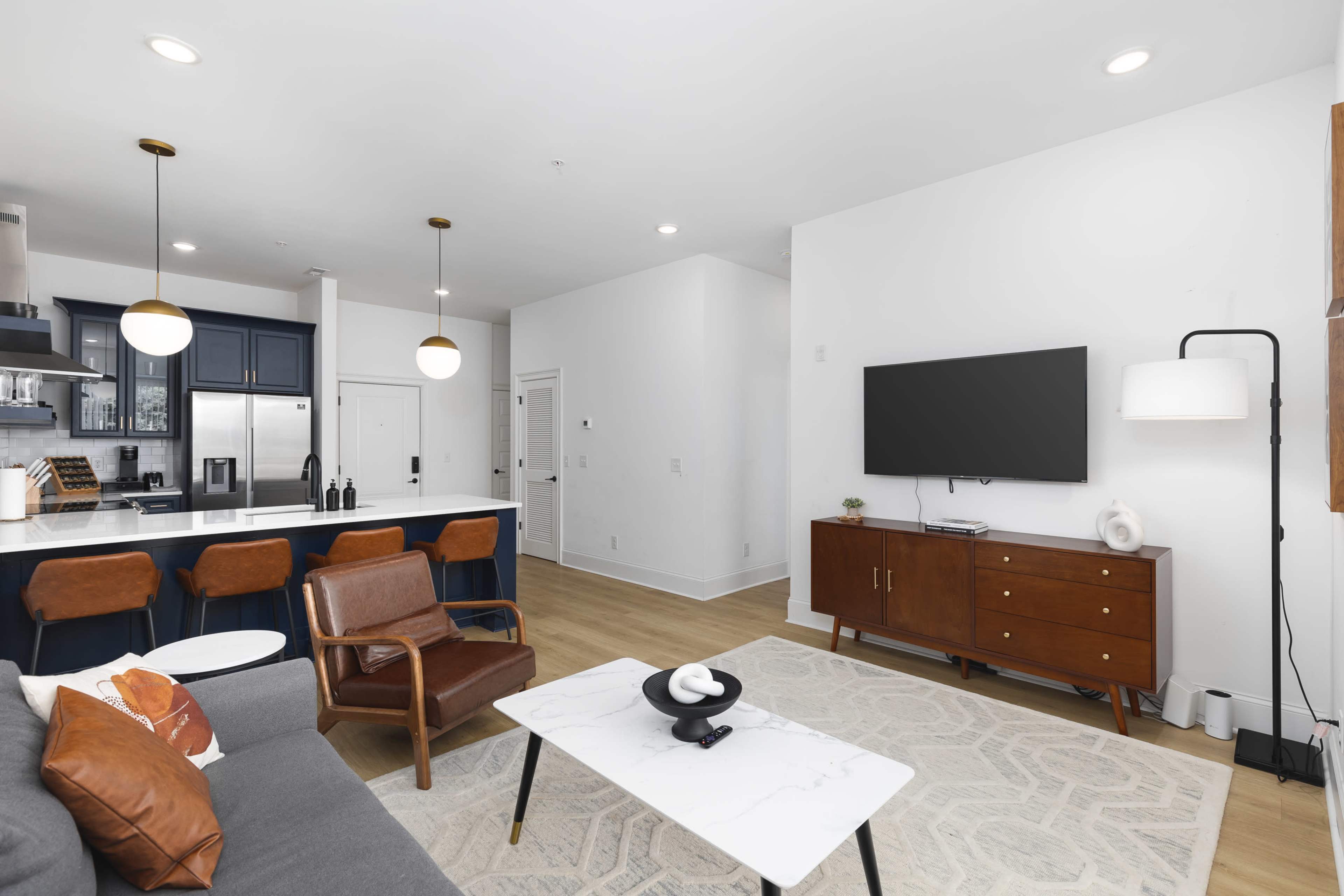 A modern living room features a gray sofa, a coffee table, a wooden sideboard, and a wall-mounted television, with a kitchen visible in the background.