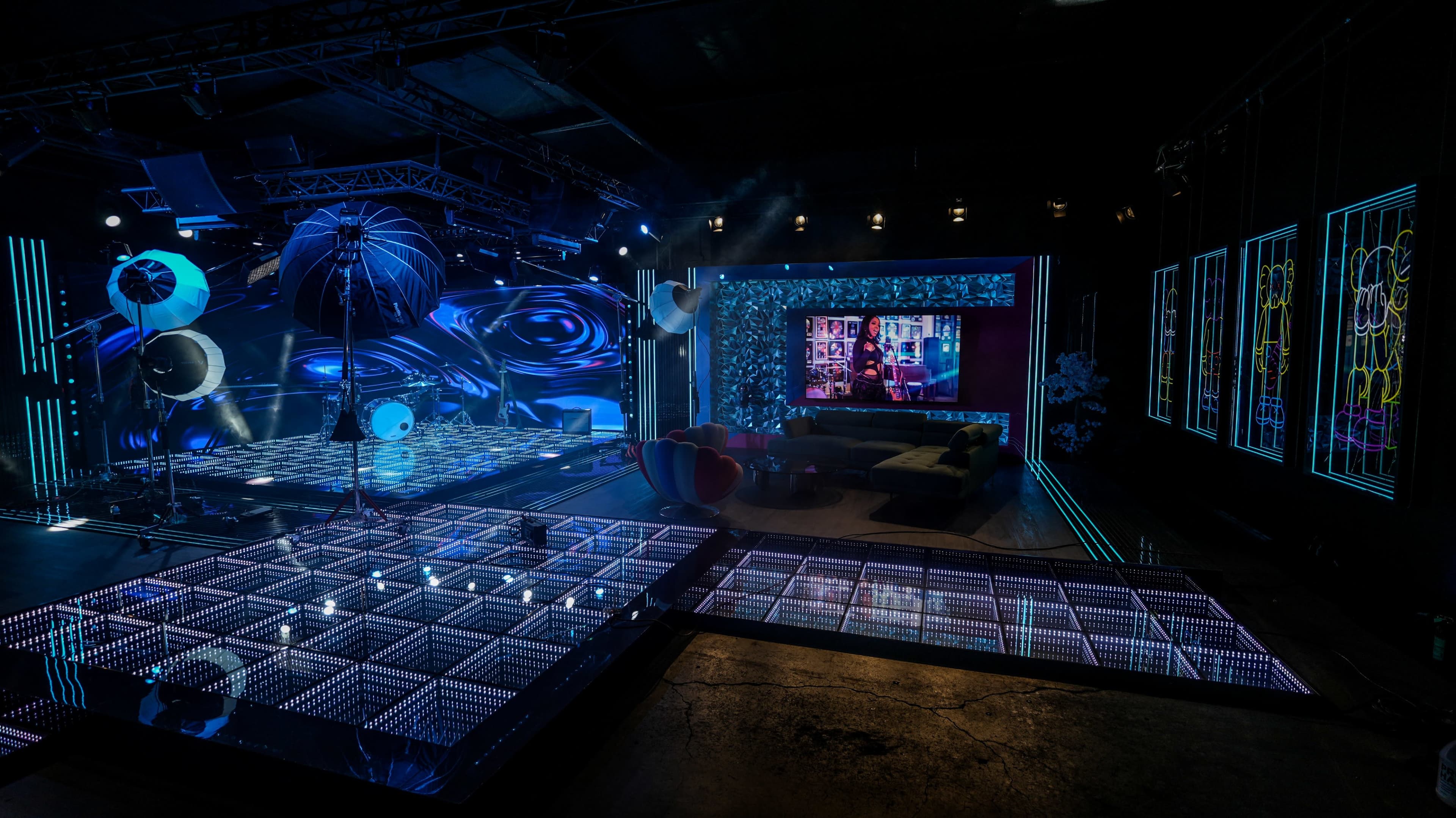 The image shows a modern television studio interior featuring a brightly lit stage with LED screens, a futuristic floor design, and a cozy seating area in the background.
