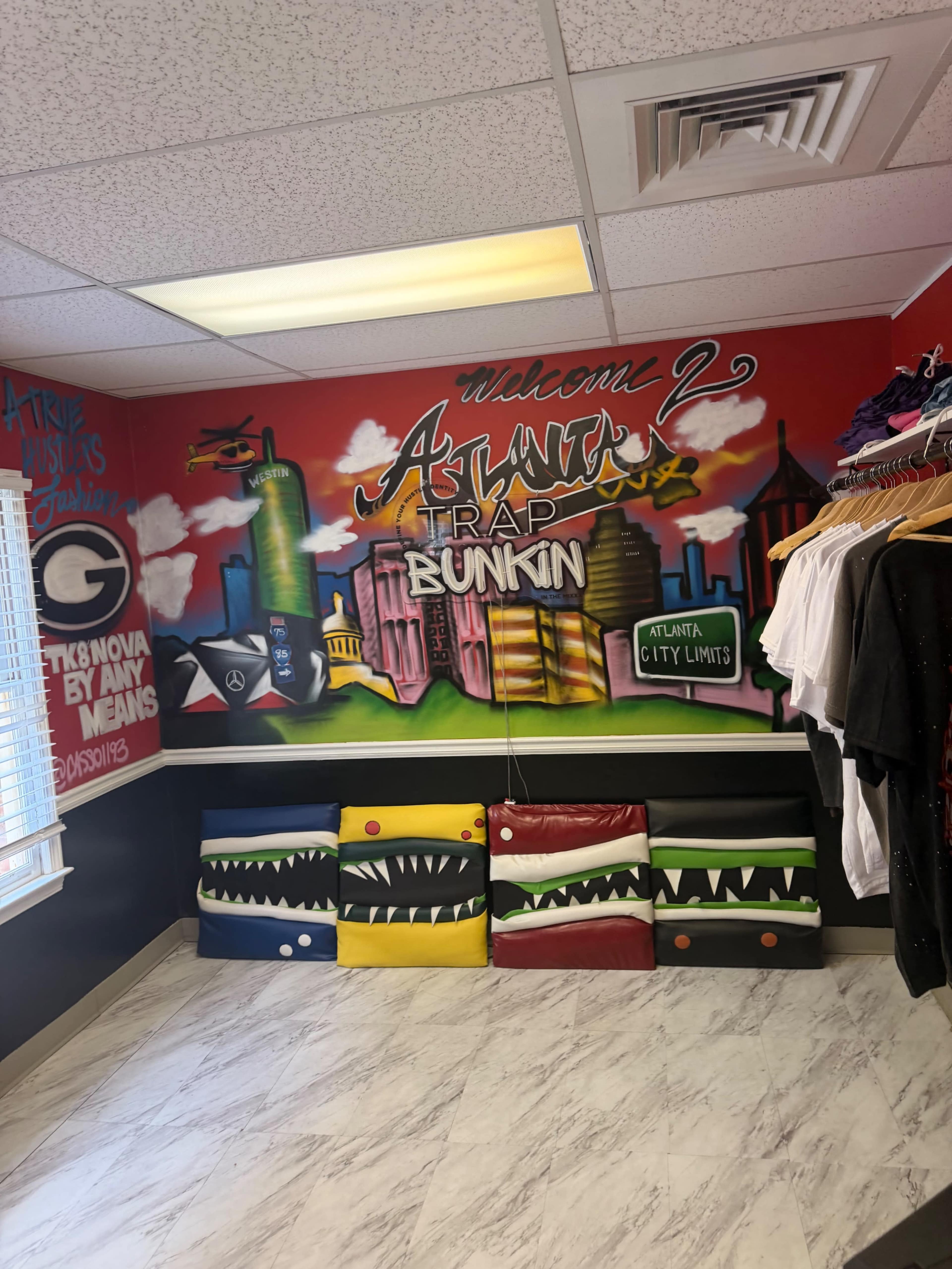 The image shows a brightly decorated room with a mural on the wall featuring a city skyline and the text "Welcome 2 Atlanta's Trap Bunkin," alongside colorful seating and clothing on display.