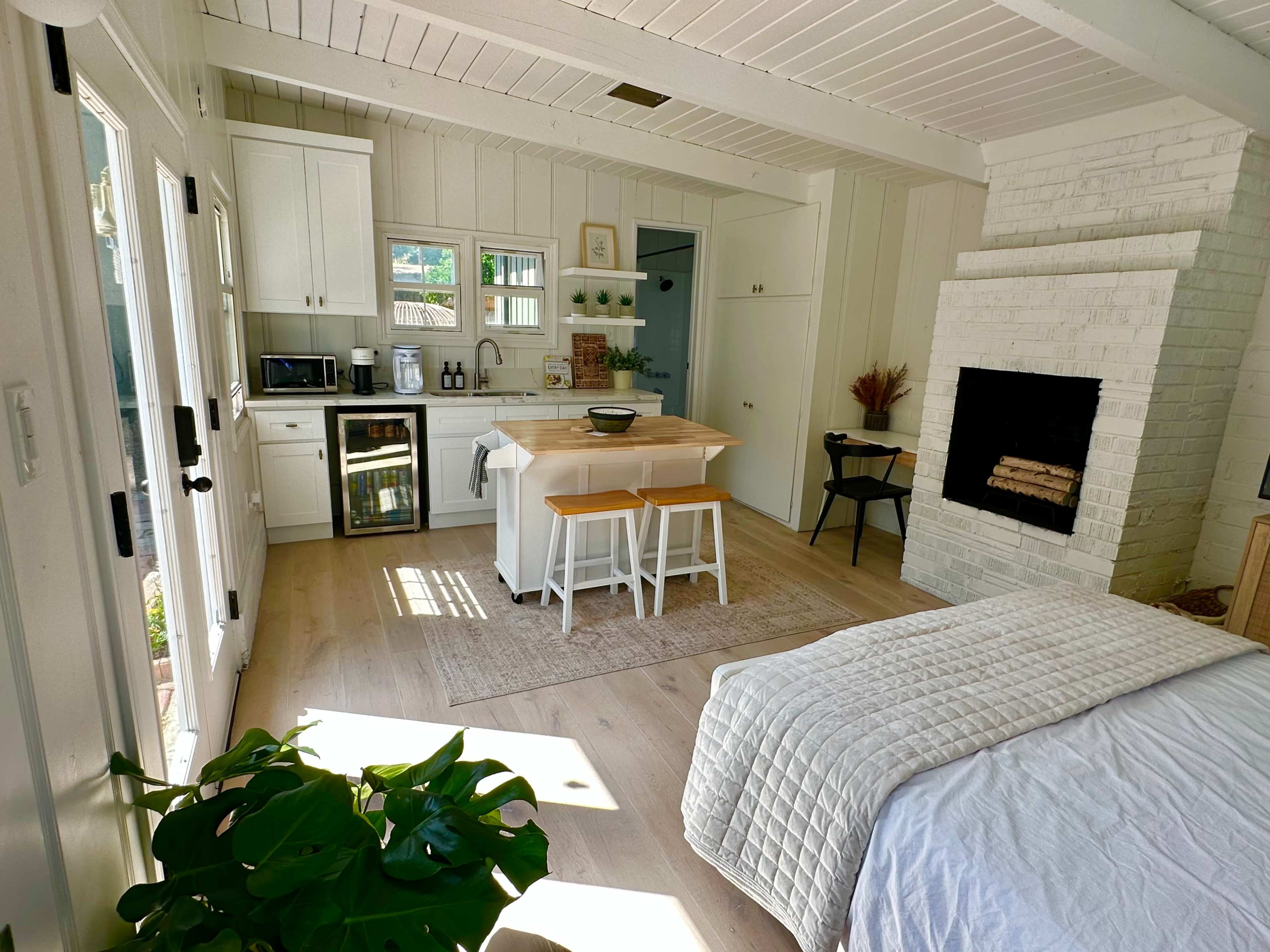 The image shows a modern, open-concept living space featuring a kitchen with white cabinets, a central island with seating, and a cozy area with a bed and a fireplace.