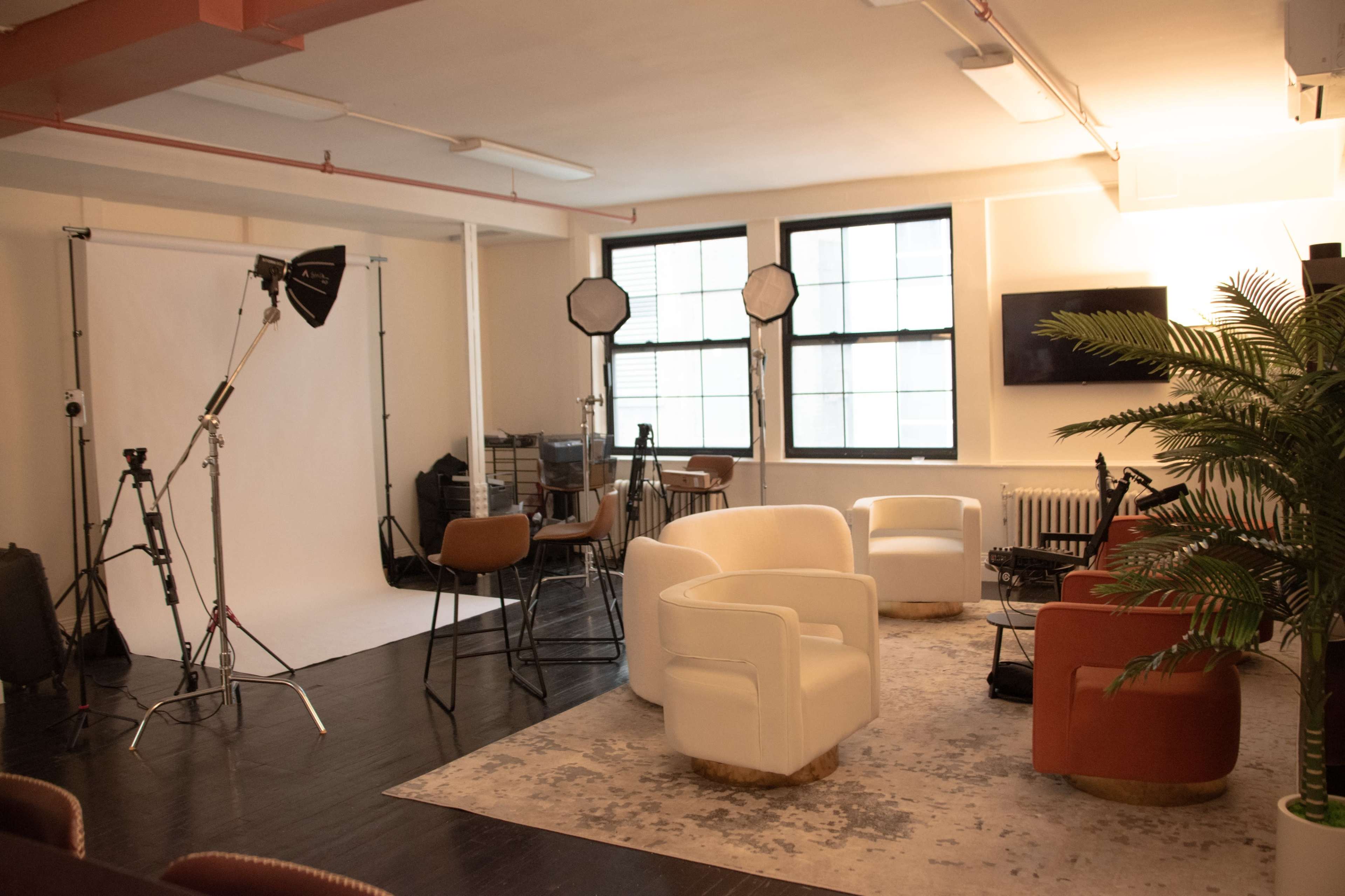 The image shows a photography studio featuring two white chairs, a red chair, various lighting equipment, and a backdrop in an open space.
