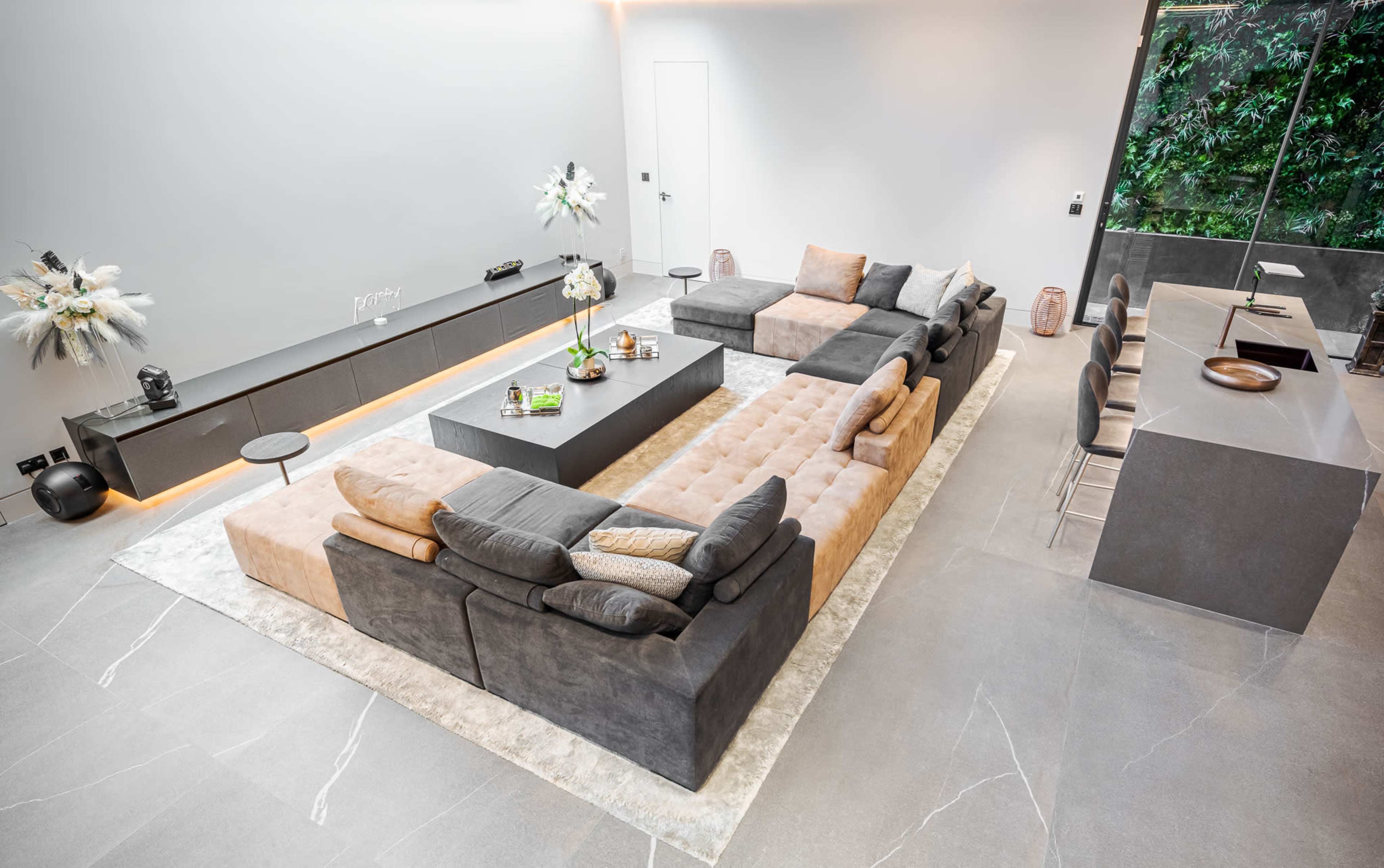 A modern open-concept living room with a large sectional sofa, a coffee table, and a dining area featuring a sleek counter and chairs.