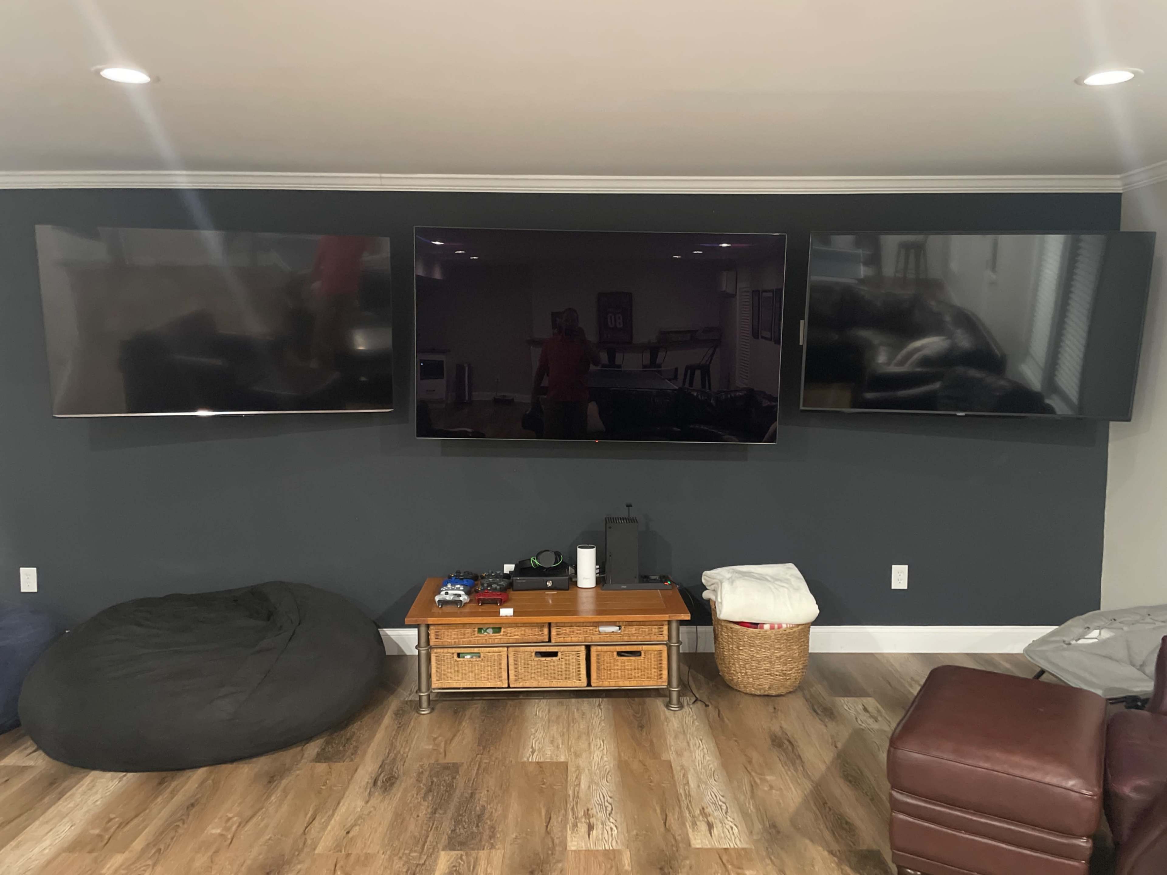 The image shows a living room wall featuring three mounted flat-screen TVs above a wooden entertainment unit and bean bag chairs.