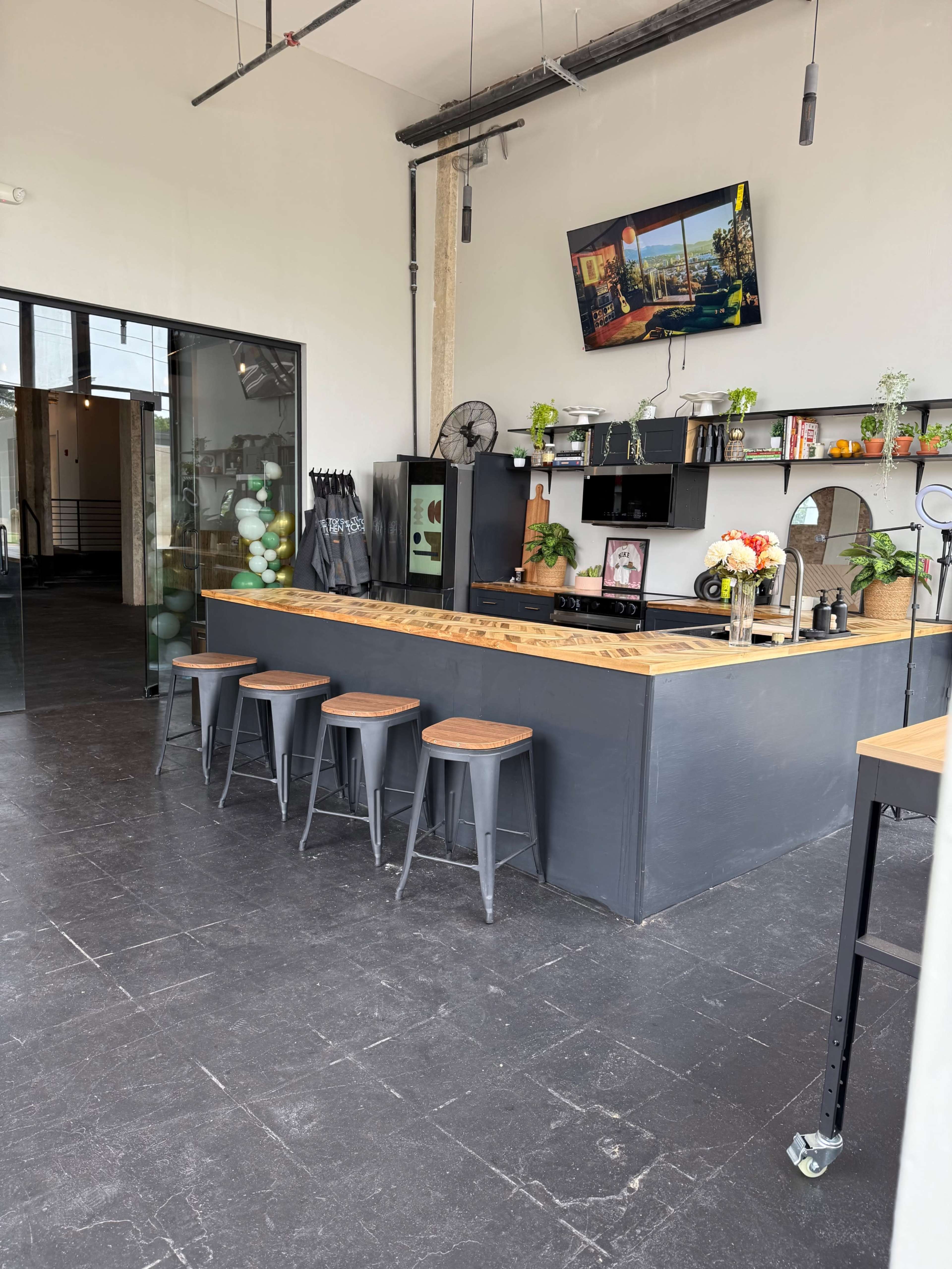 The image shows a modern café interior with a wooden bar, industrial-style stools, and decorative plants, along with a wall-mounted television.