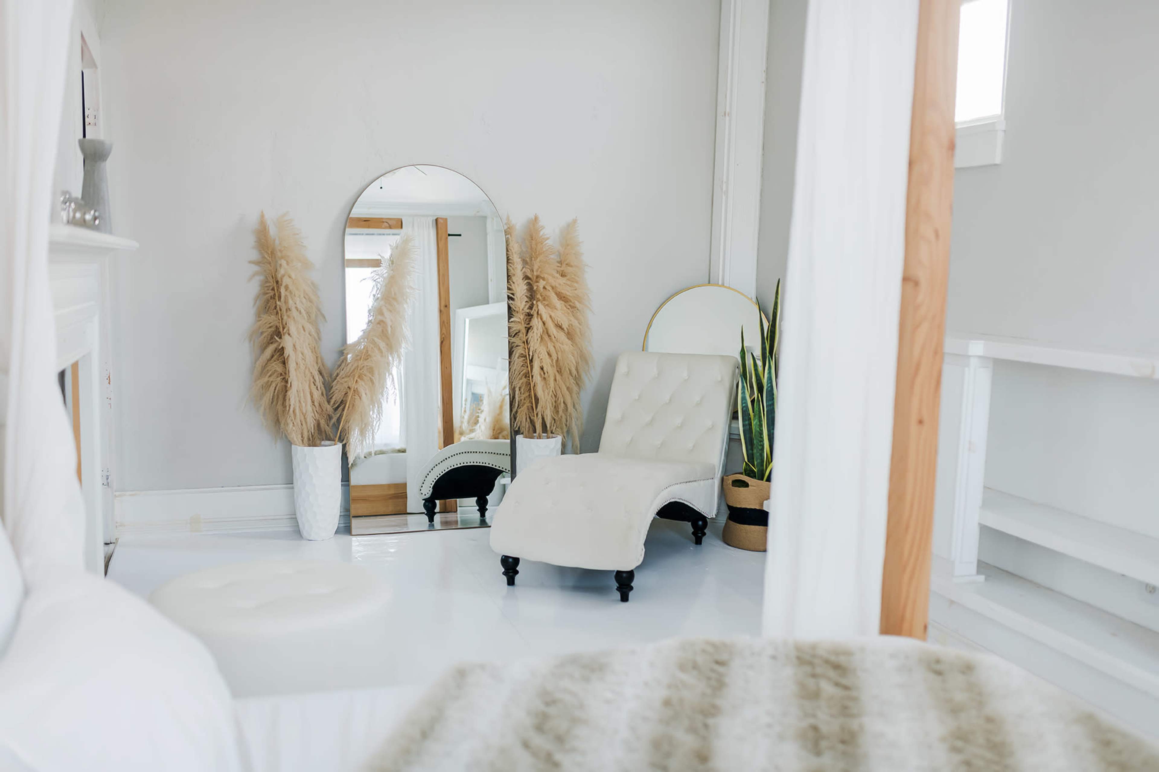 The image shows a bright, minimalist bedroom featuring a white chaise lounge, large mirror, and decorative plants in neutral-colored pots.