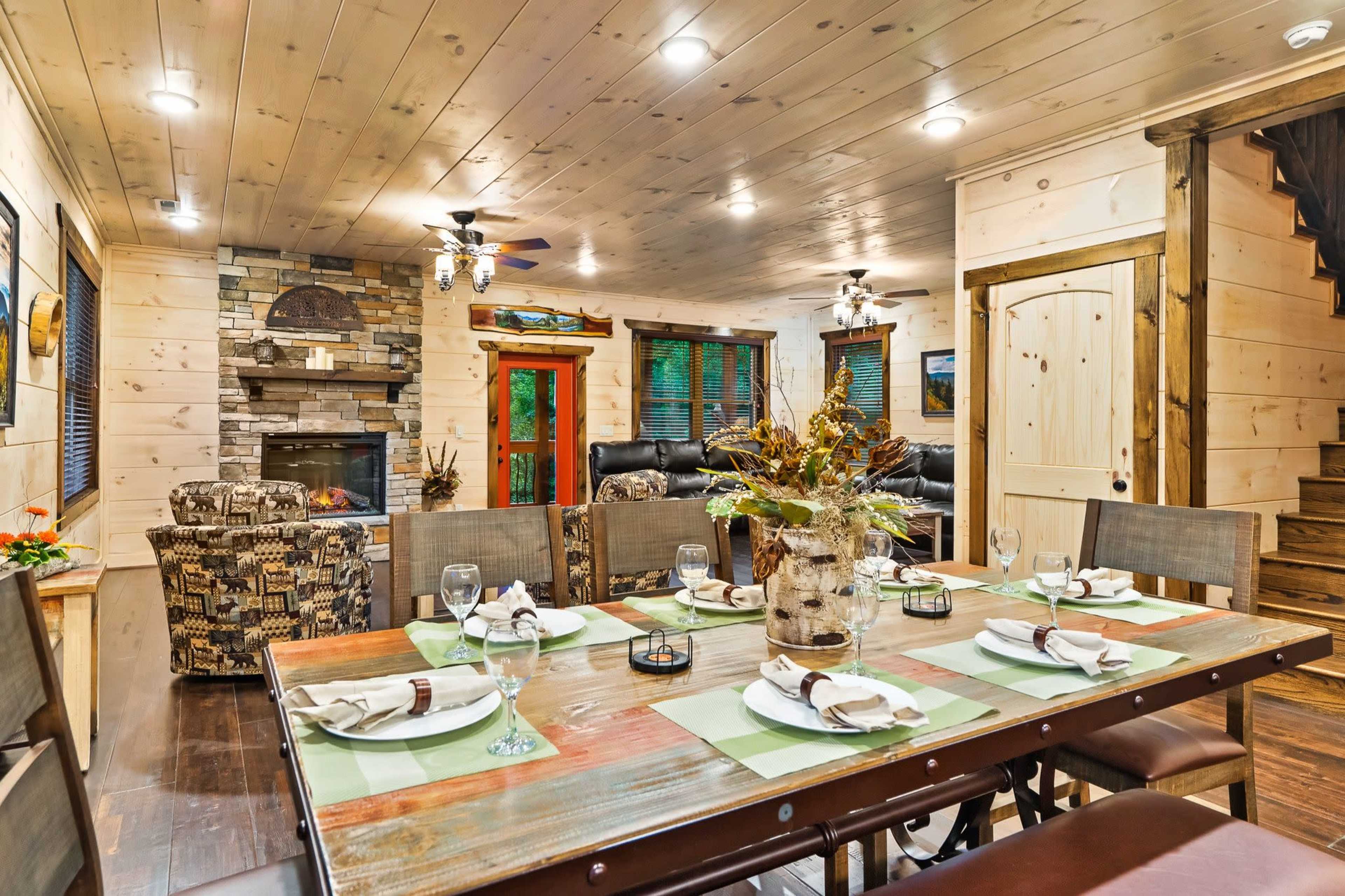 A dining table is set for a meal in a cozy wooden-paneled room featuring a stone fireplace and a staircase.