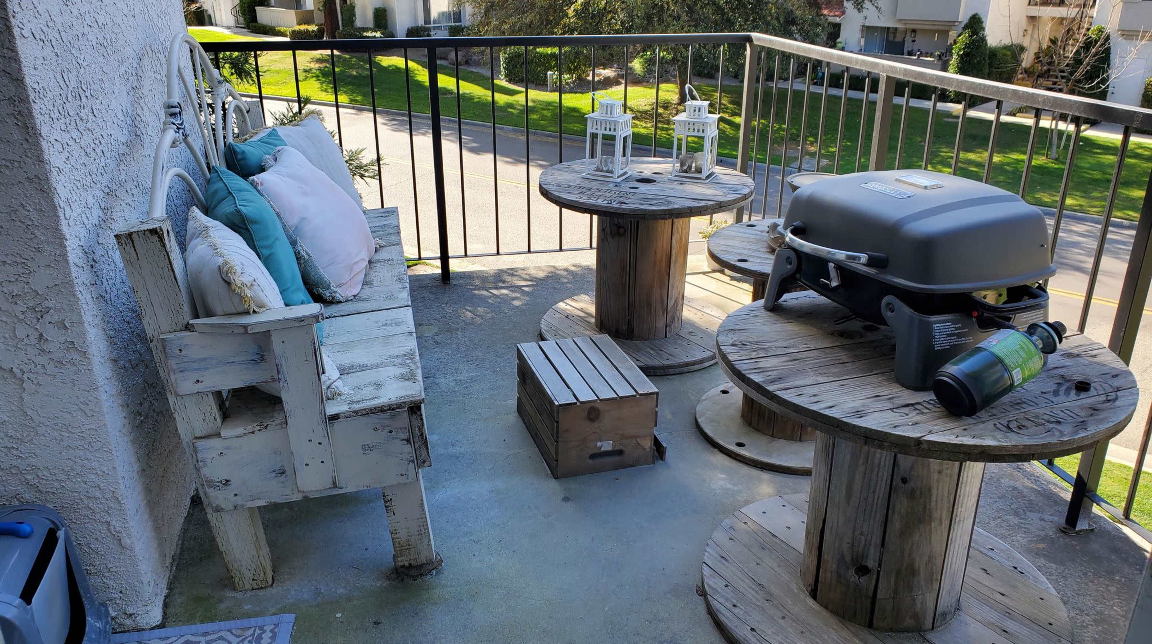A balcony with a wooden bench, two round tables made from pallets, a portable grill, and decorative lanterns.