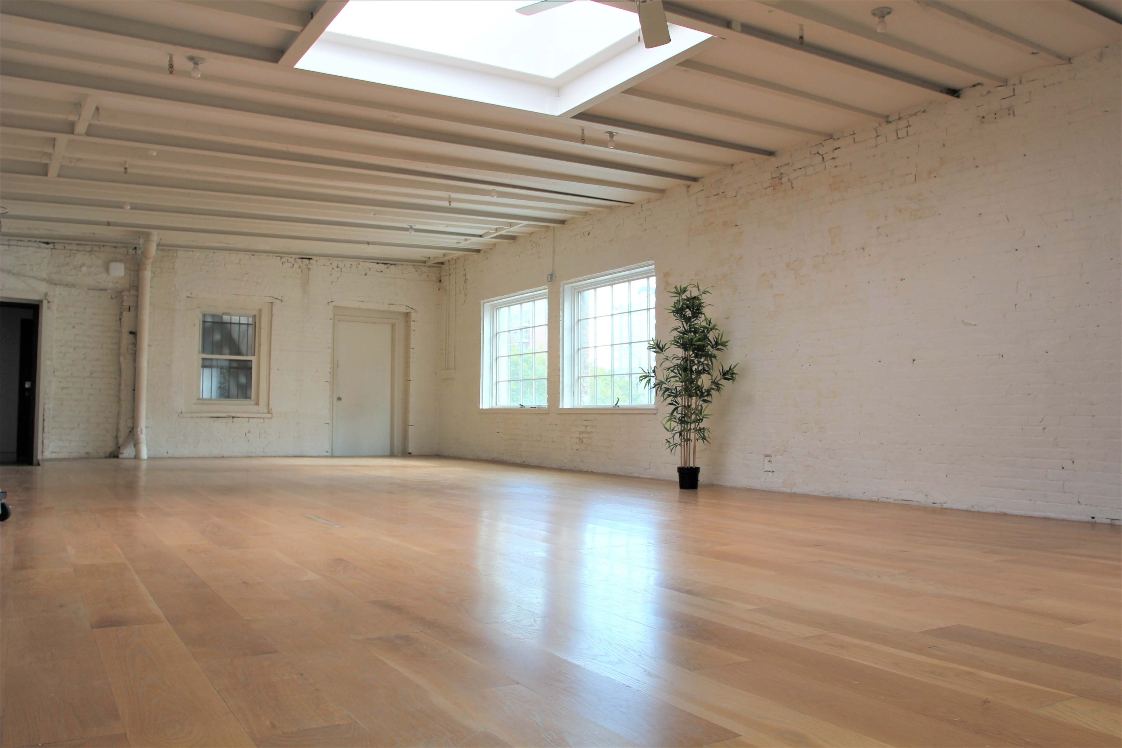 The image shows a spacious, bright room with wooden flooring, white brick walls, and large windows, featuring a small potted plant in one corner.