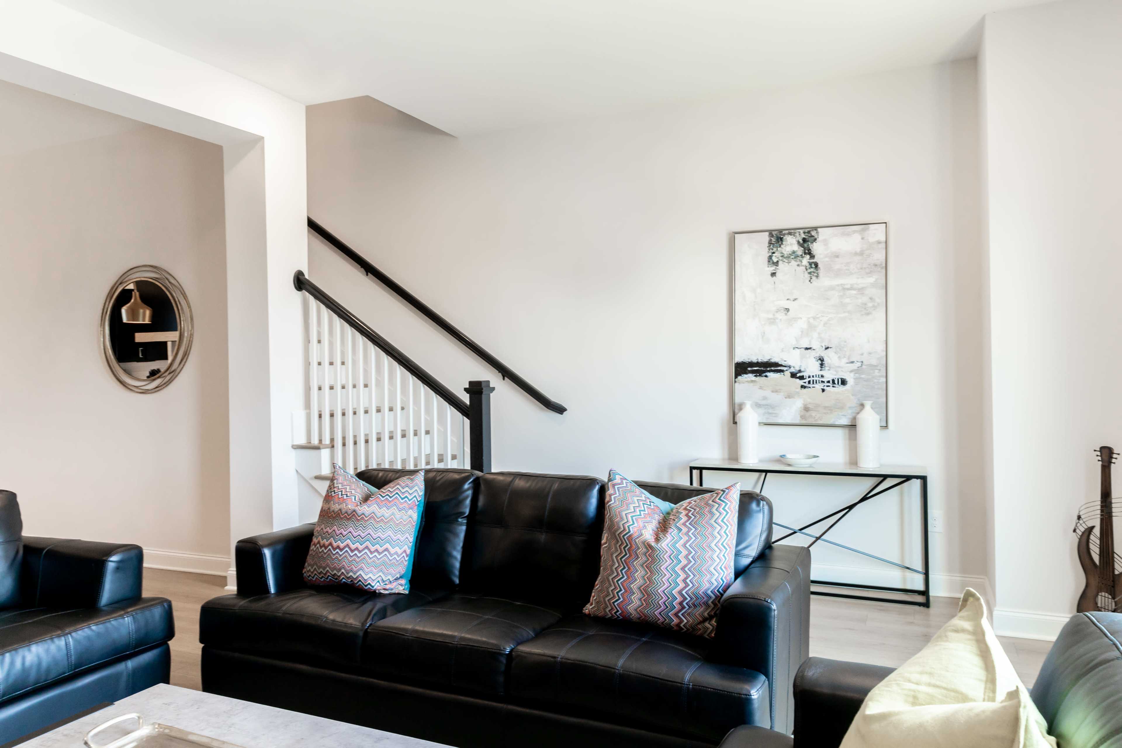 A living room features black leather sofas with patterned throw pillows, a wall-mounted circular mirror, and a staircase in the background.