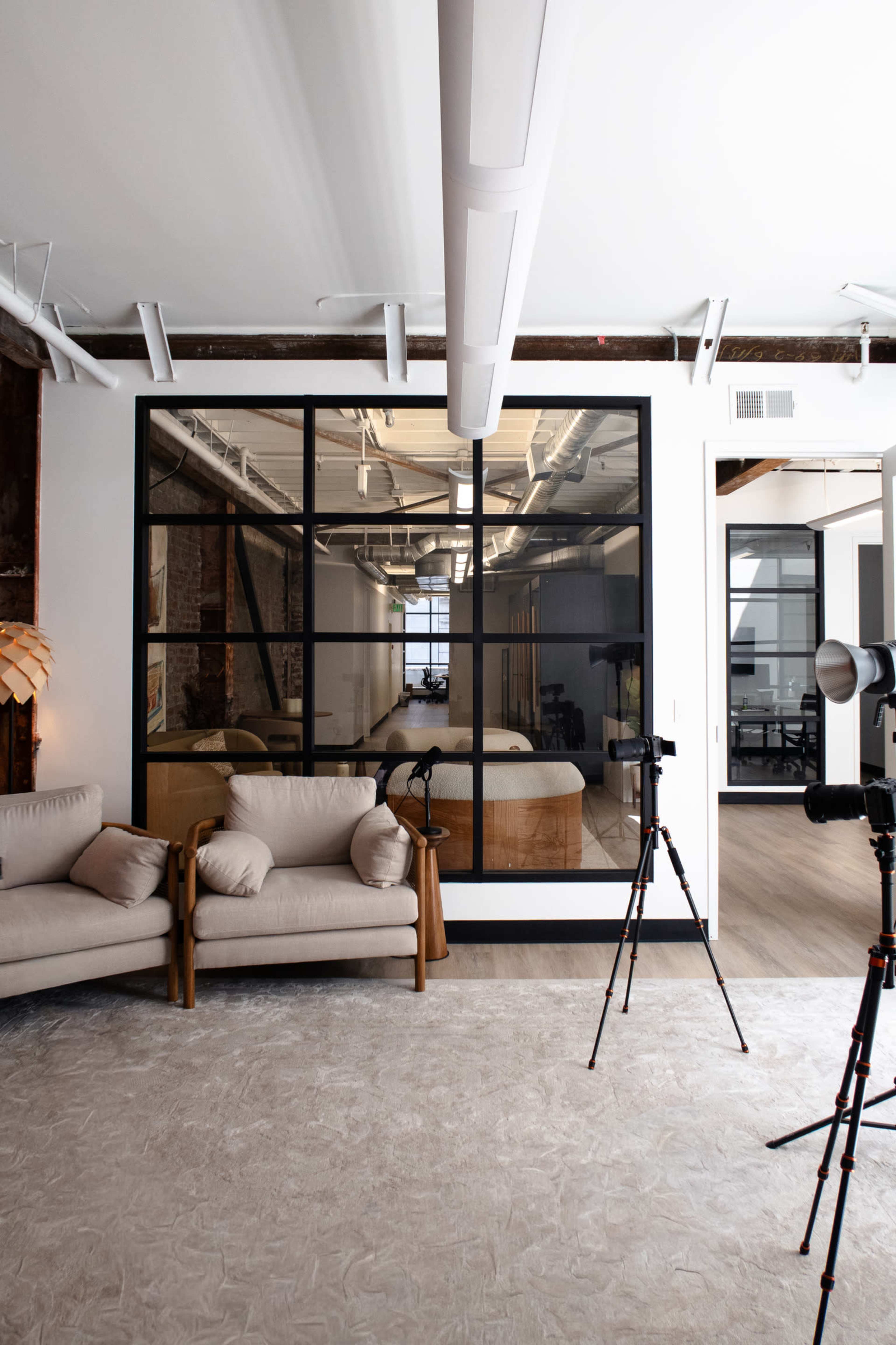 The image shows a modern office space featuring a seating area with light-colored sofas, a large glass partition, and photography equipment positioned on a gray rug.