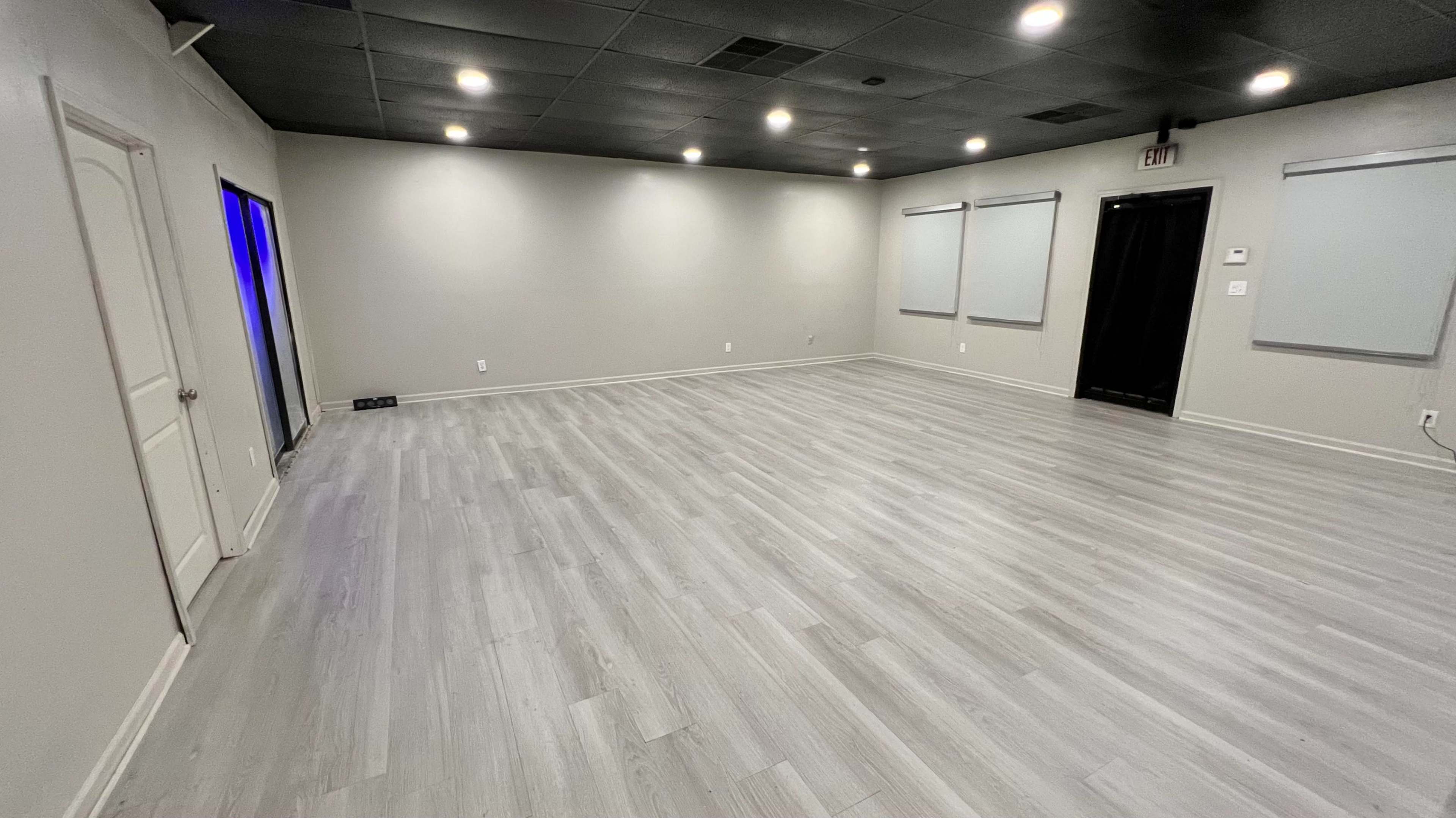 The image shows a spacious, empty room with gray walls and light-colored wooden flooring, featuring several overhead lights and a door on the left.