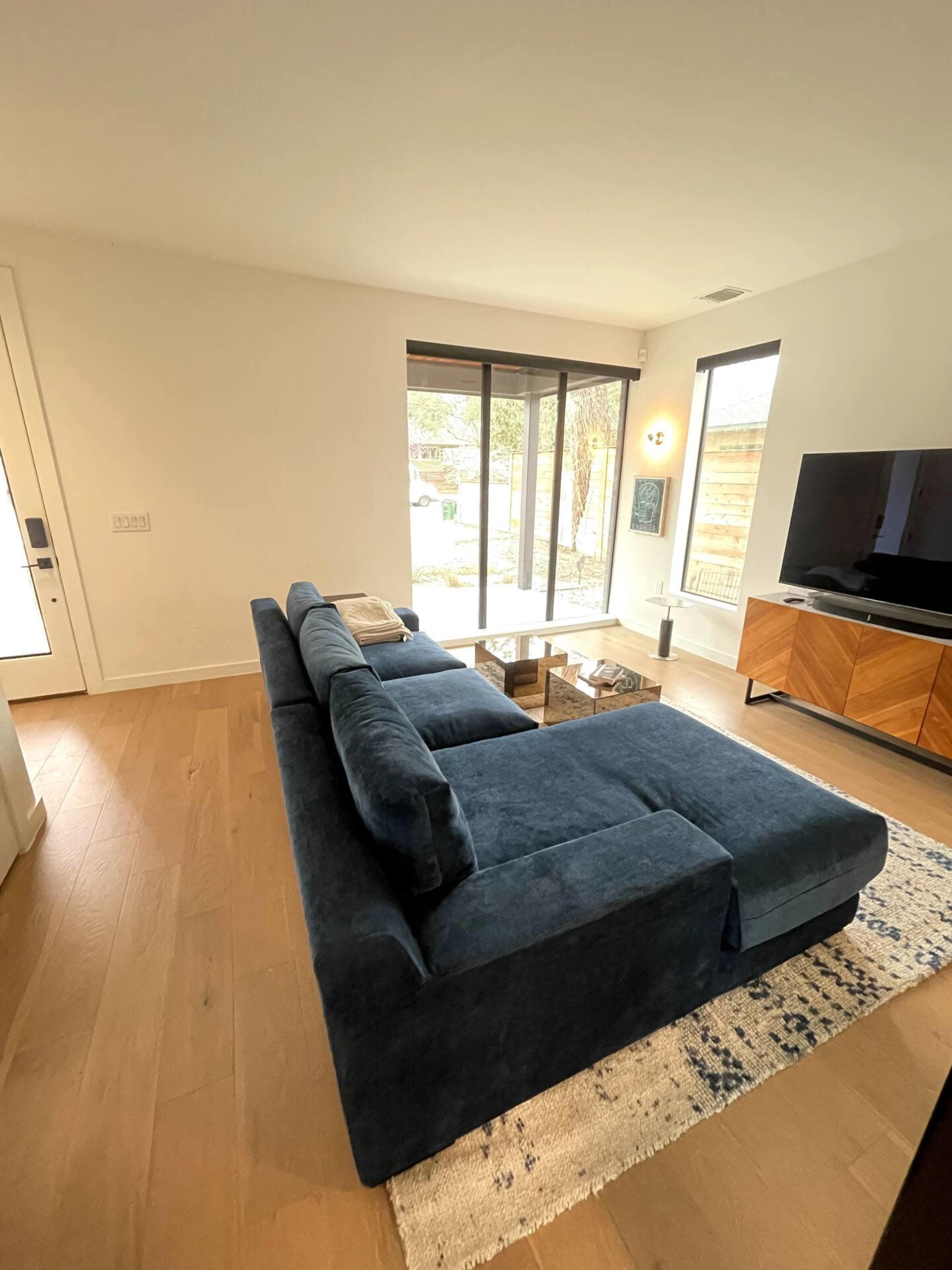 A dark blue sectional couch is positioned near a large window, facing a wooden TV stand with a flat-screen television in a modern living room.