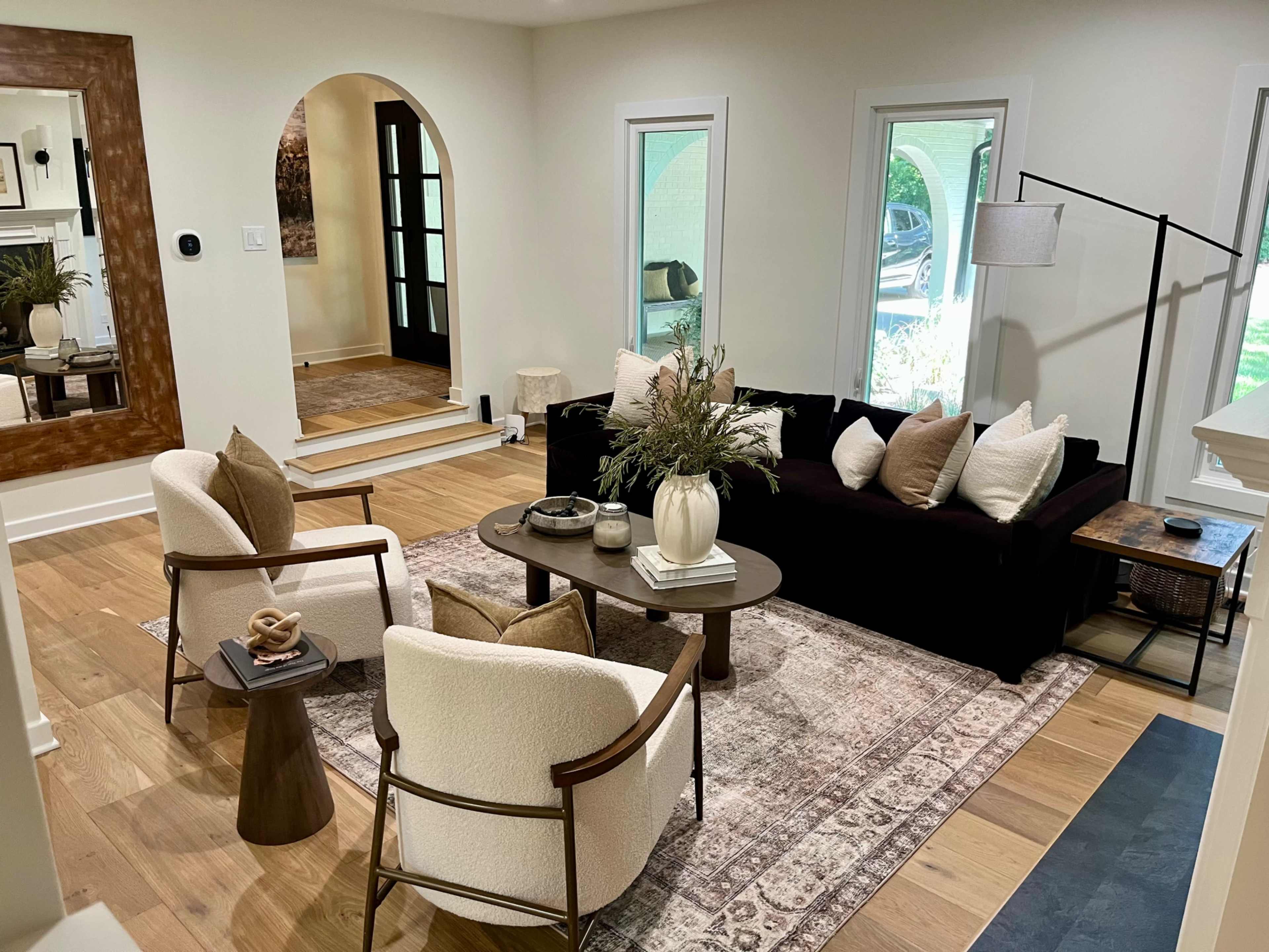 The image shows a modern living room featuring a black sectional sofa, two upholstered chairs, a round coffee table, and large windows providing natural light.