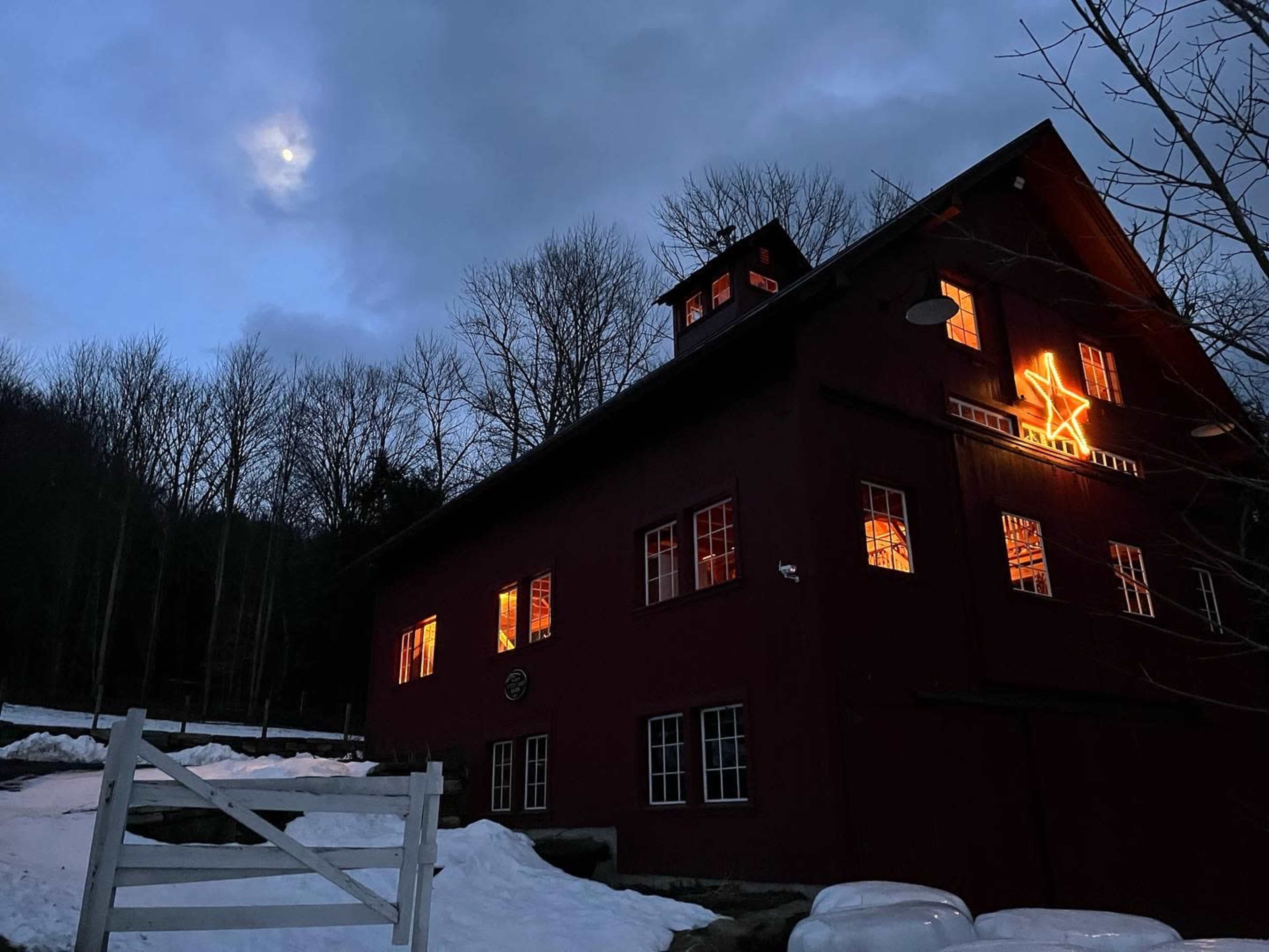 A red wooden house is illuminated at dusk with a star decoration in the window, surrounded by snow and bare trees beneath a partially visible moon.