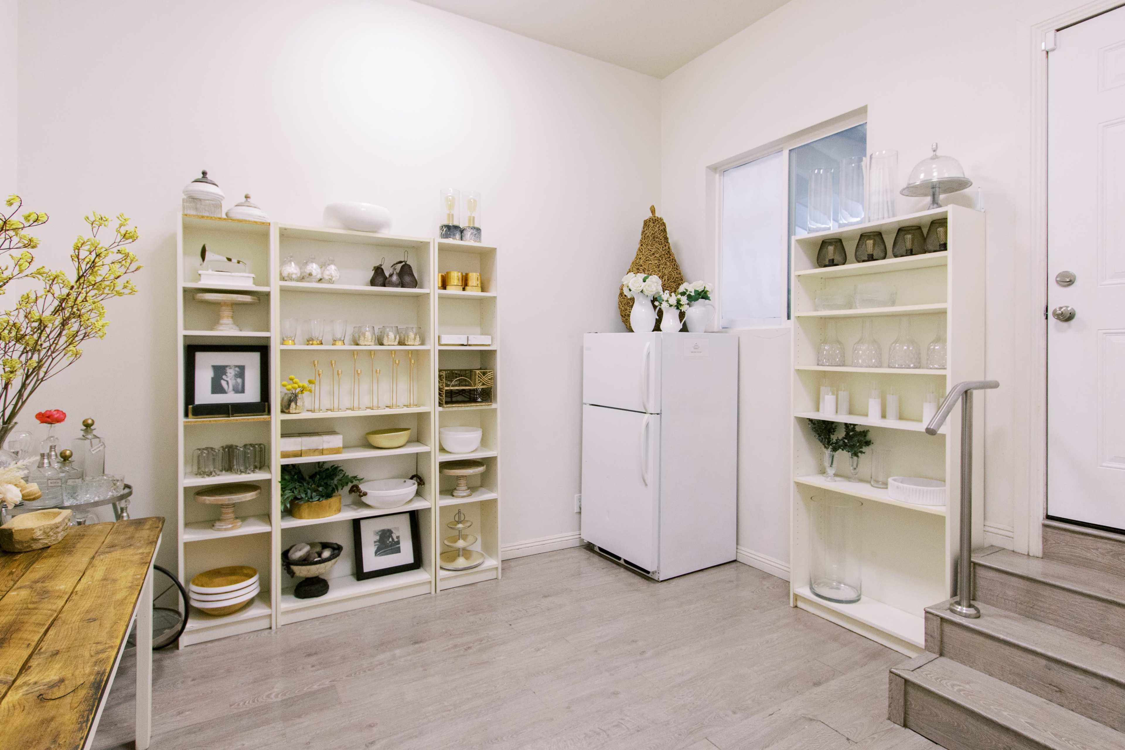 A well-organized room with white shelves displaying various dishes and decor, a white refrigerator, and a door leading outside.