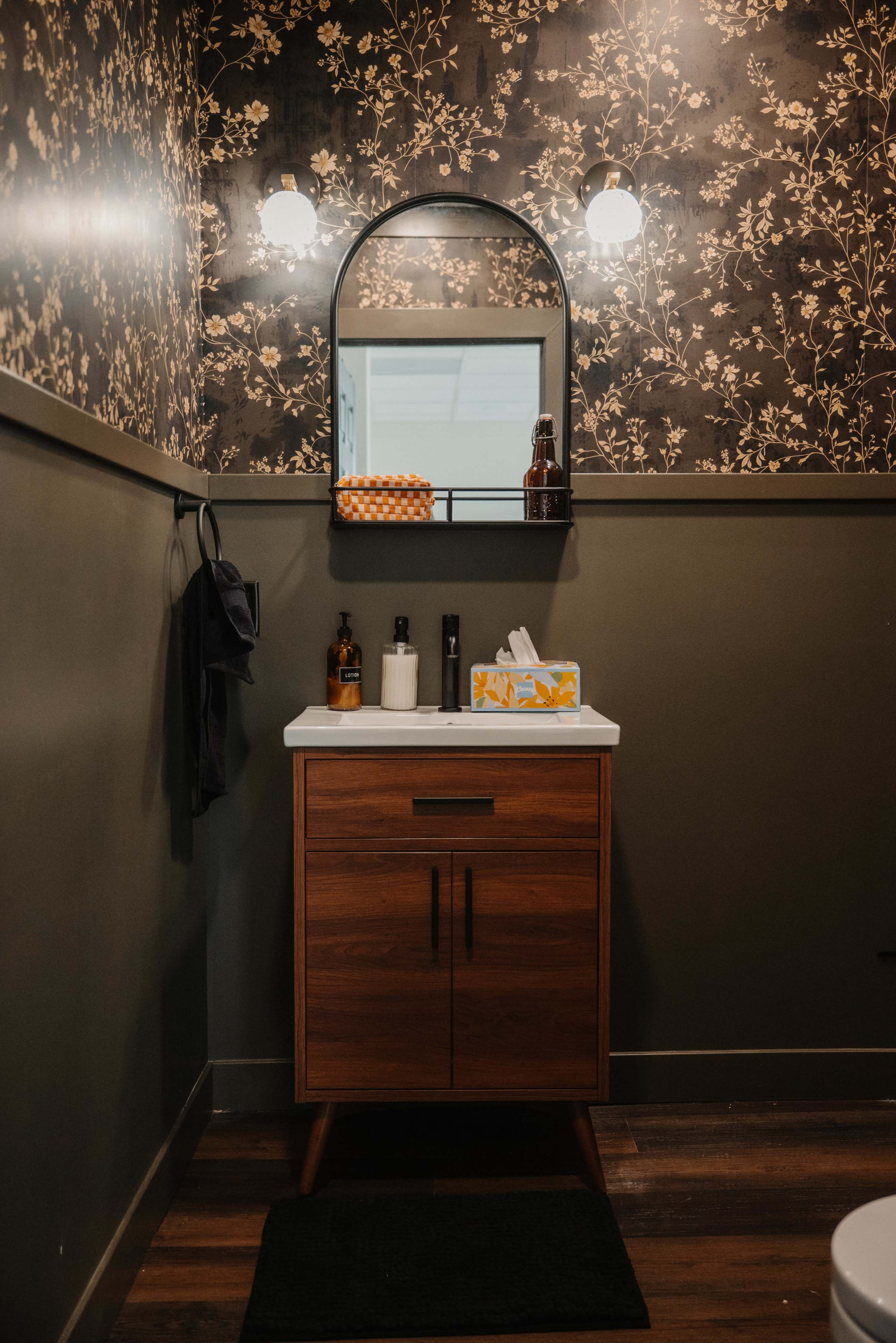 The image shows a small bathroom featuring a wooden vanity with a white countertop, a mirror with an arched frame above it, and a wall adorned with floral-patterned wallpaper.