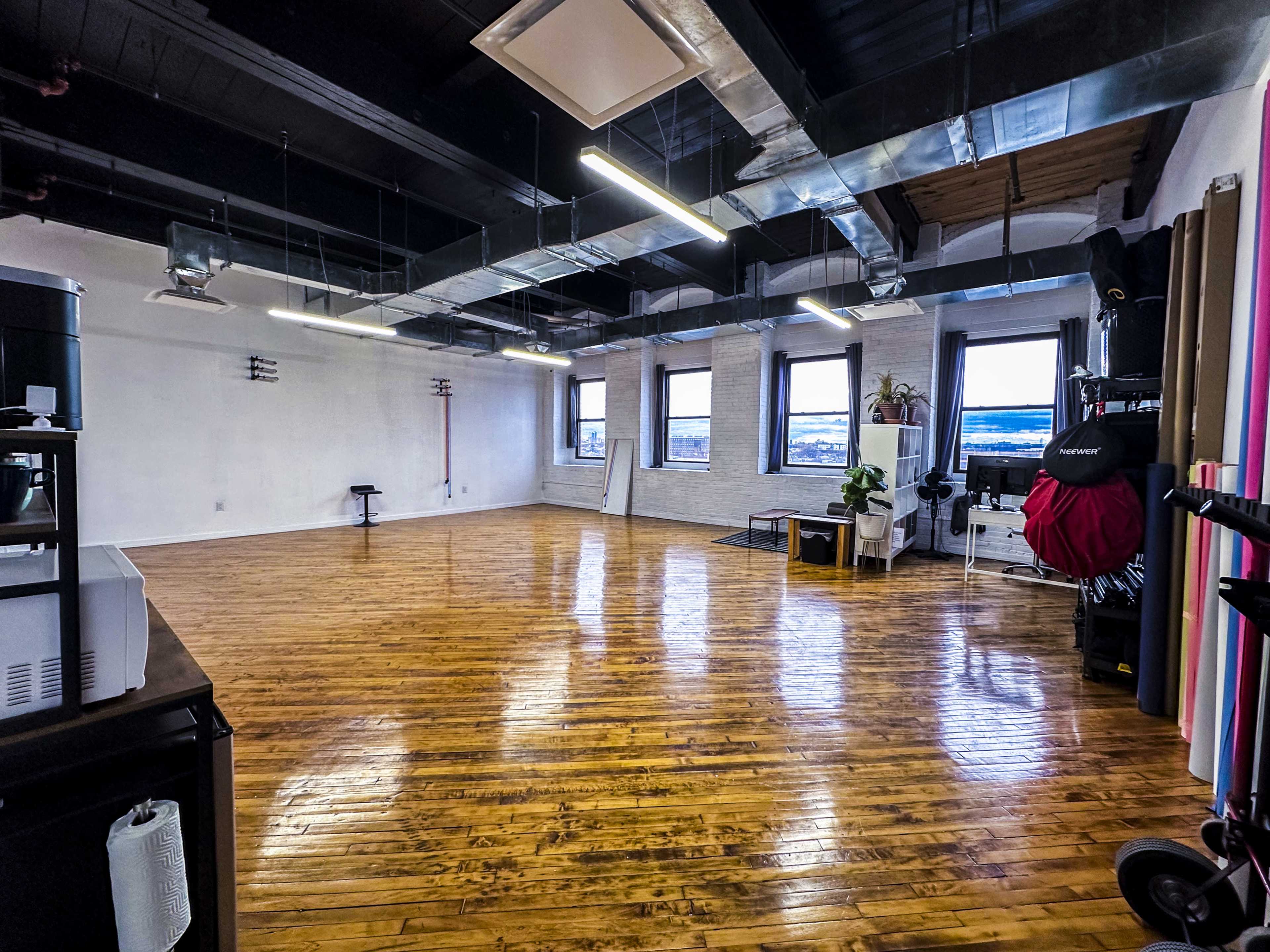 A spacious, empty room with polished wooden floors, large windows, and exposed ductwork.
