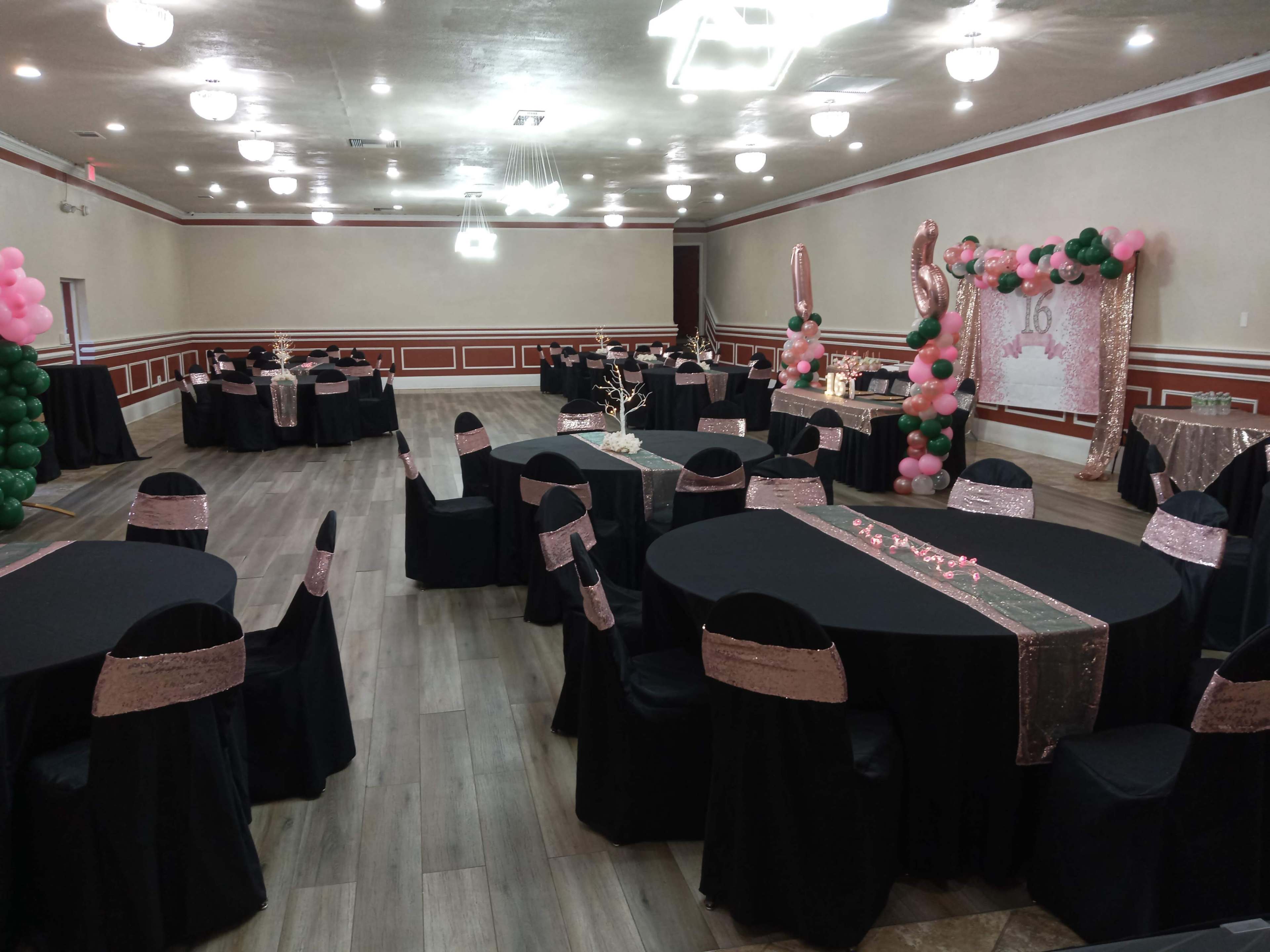 The image shows a decorated event space featuring multiple round black tables with black chairs and pink accents, including balloons and a backdrop for a celebration.