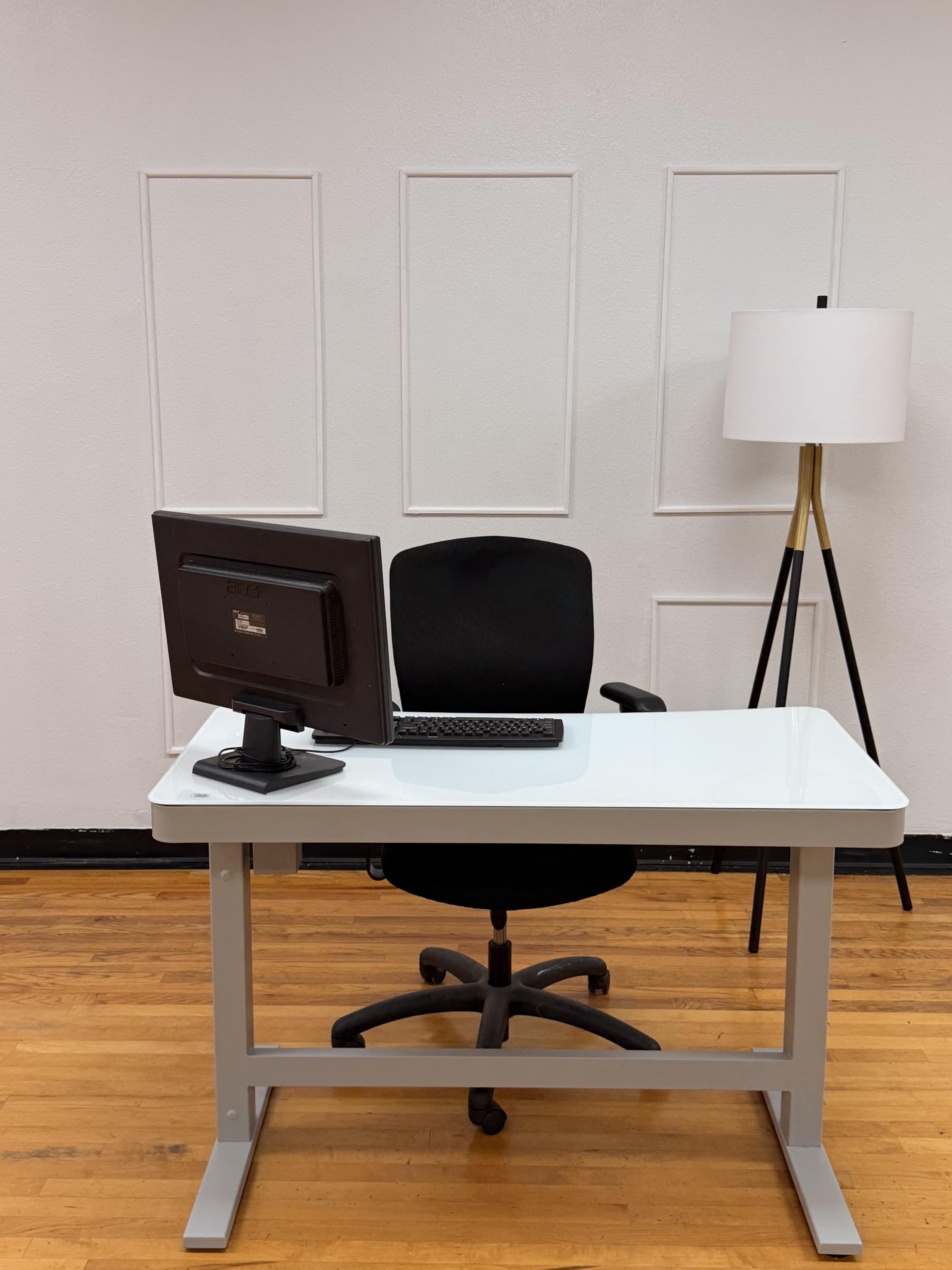 The image shows a minimalistic office setup featuring a computer on a white desk, a black office chair, and a standing lamp against a plain wall.
