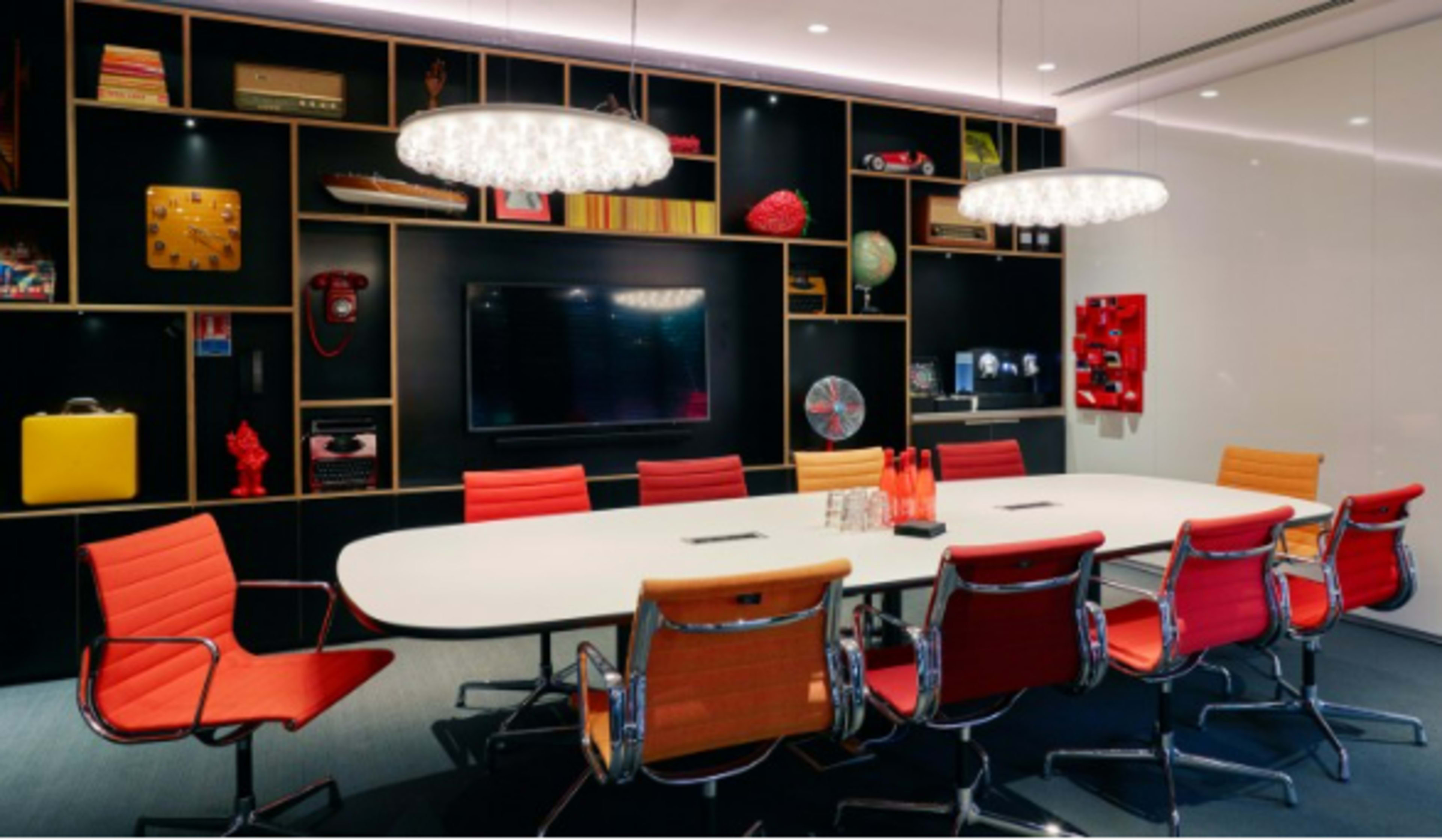 A modern conference room with a large white table surrounded by red and orange chairs, featuring decorative shelves with various items and a television mounted on the wall.