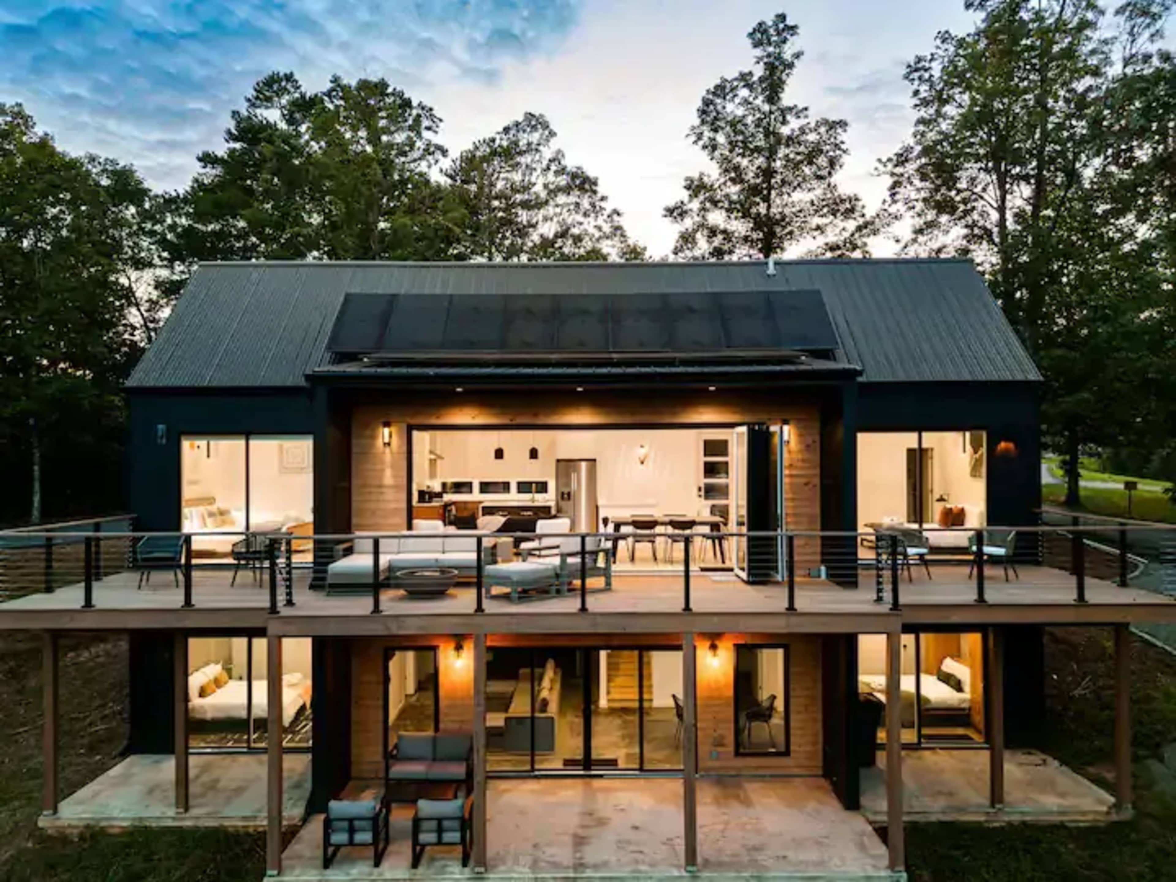 A modern two-story house with large windows and a spacious wooden deck surrounded by trees at dusk.