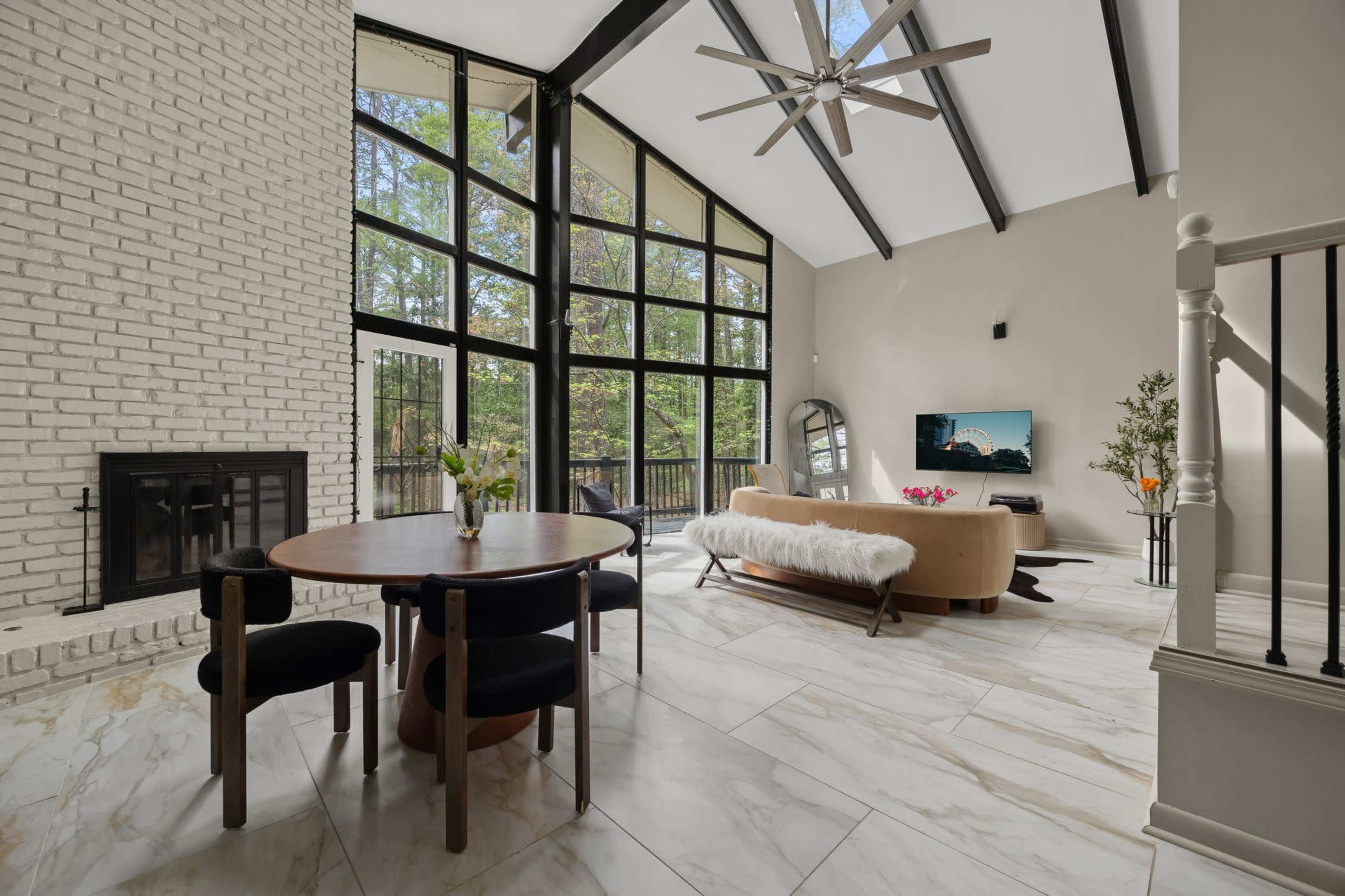 The image features a modern living space with large windows, a round dining table, a sofa, and a fireplace, accentuated by high ceilings and visible beams.