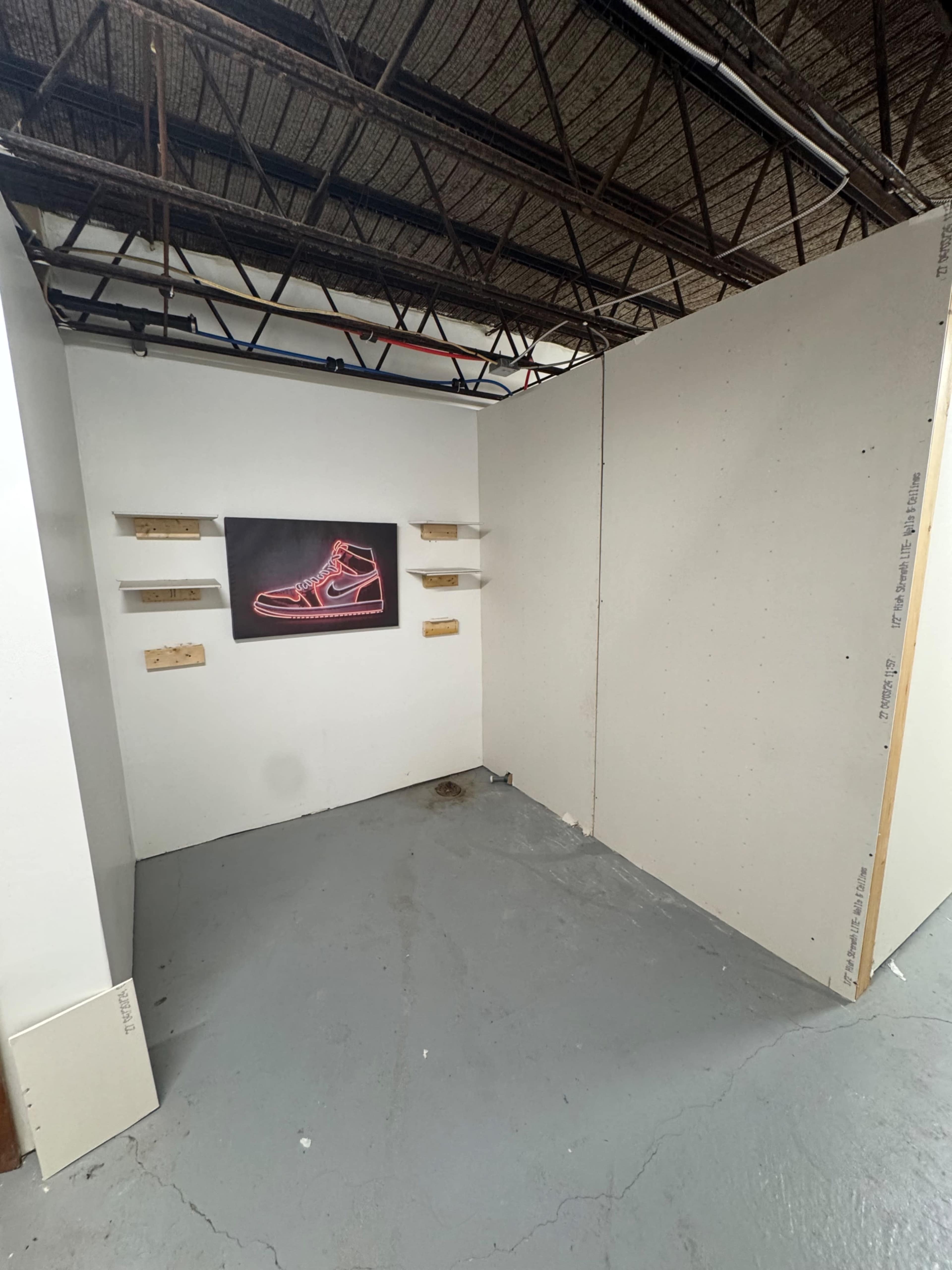 The image shows an empty room with exposed walls, featuring a vibrant poster of a sneaker on one wall and three wooden shelves mounted on the adjacent wall.