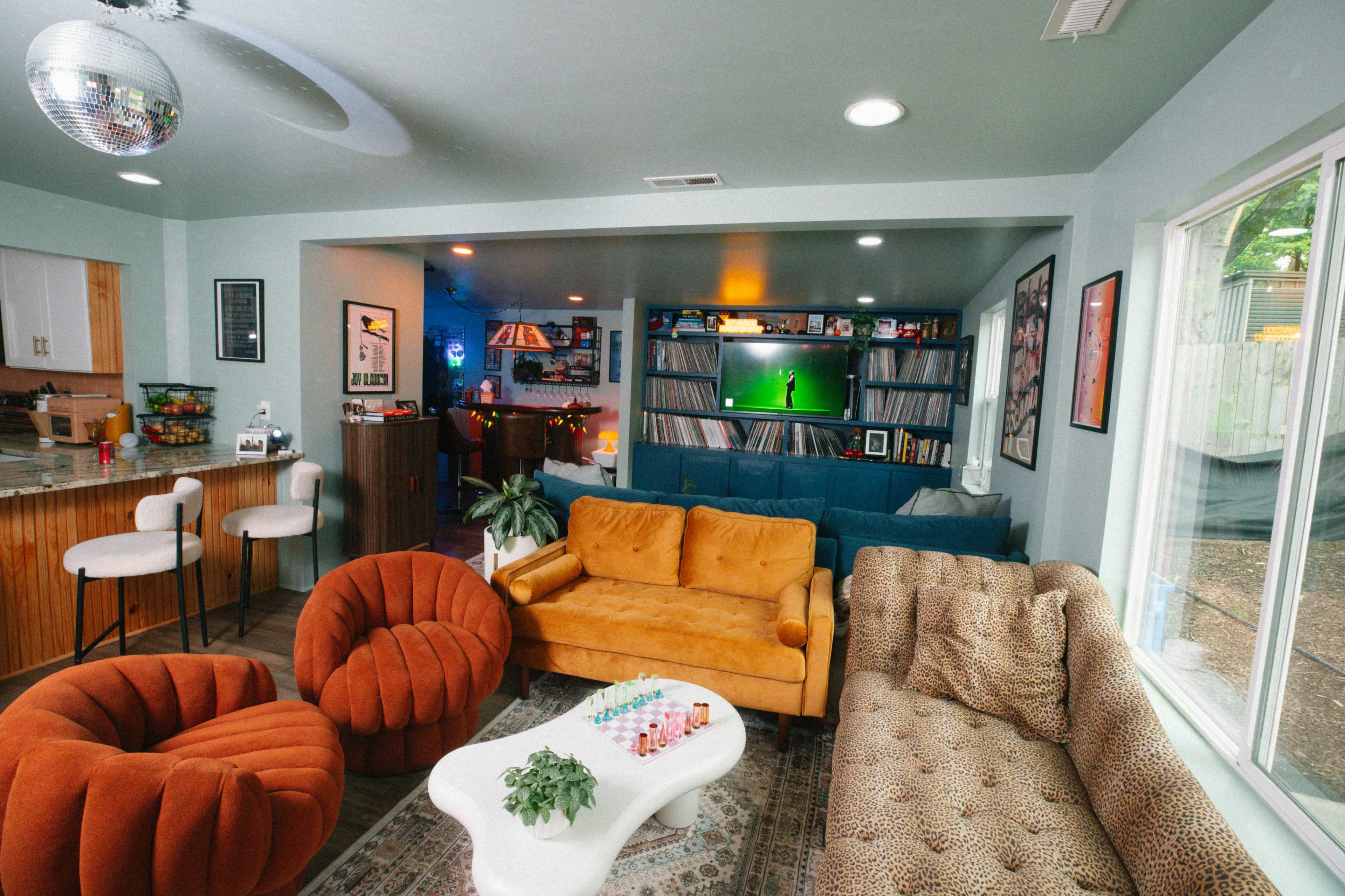 The image shows a retro-styled living room featuring a mix of colorful furniture, shelves of records, and a disco ball, with a cozy vibe enhanced by warm lighting.