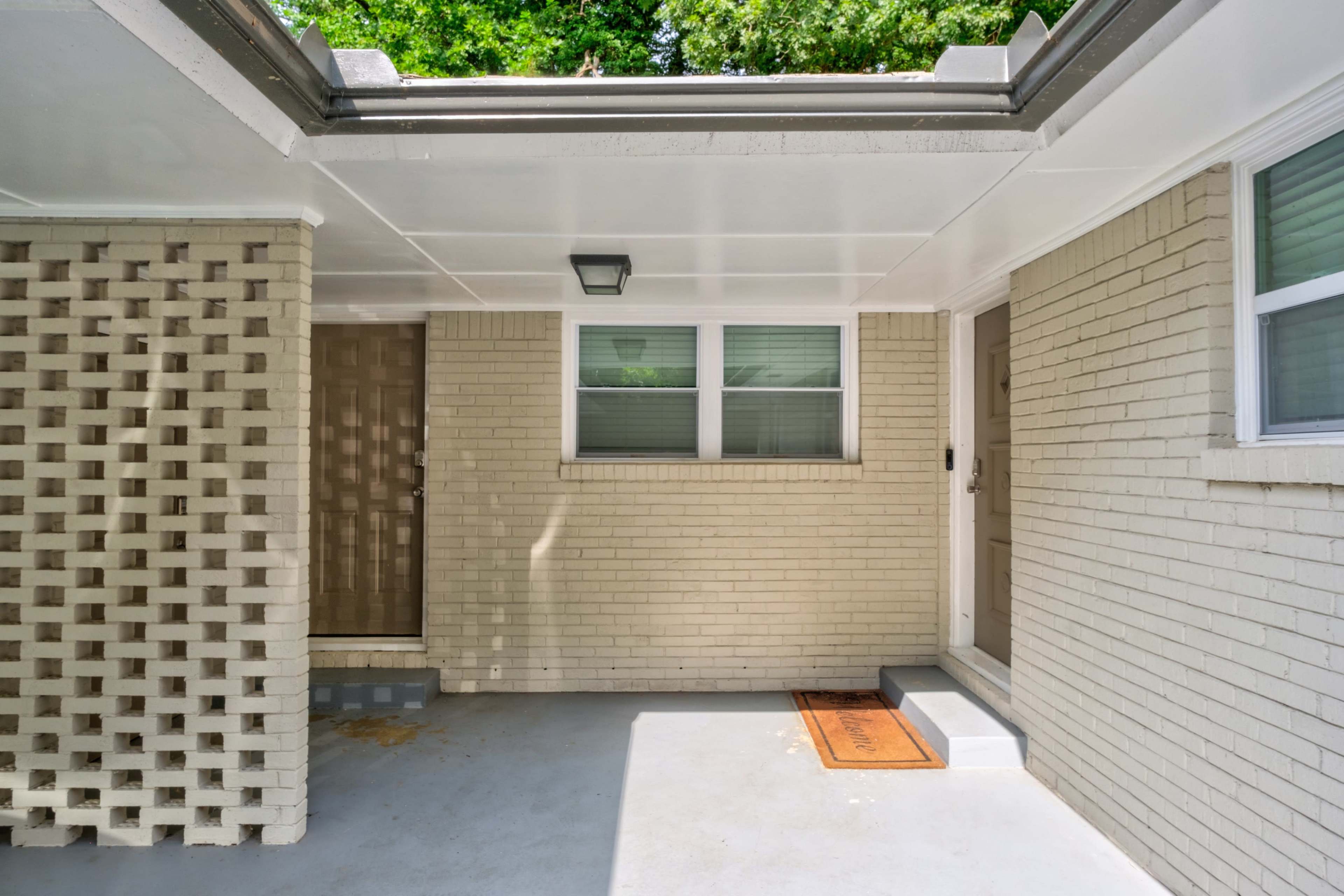 A covered front porch area with a beige brick wall, a door with a window, and a welcome mat at the entrance.