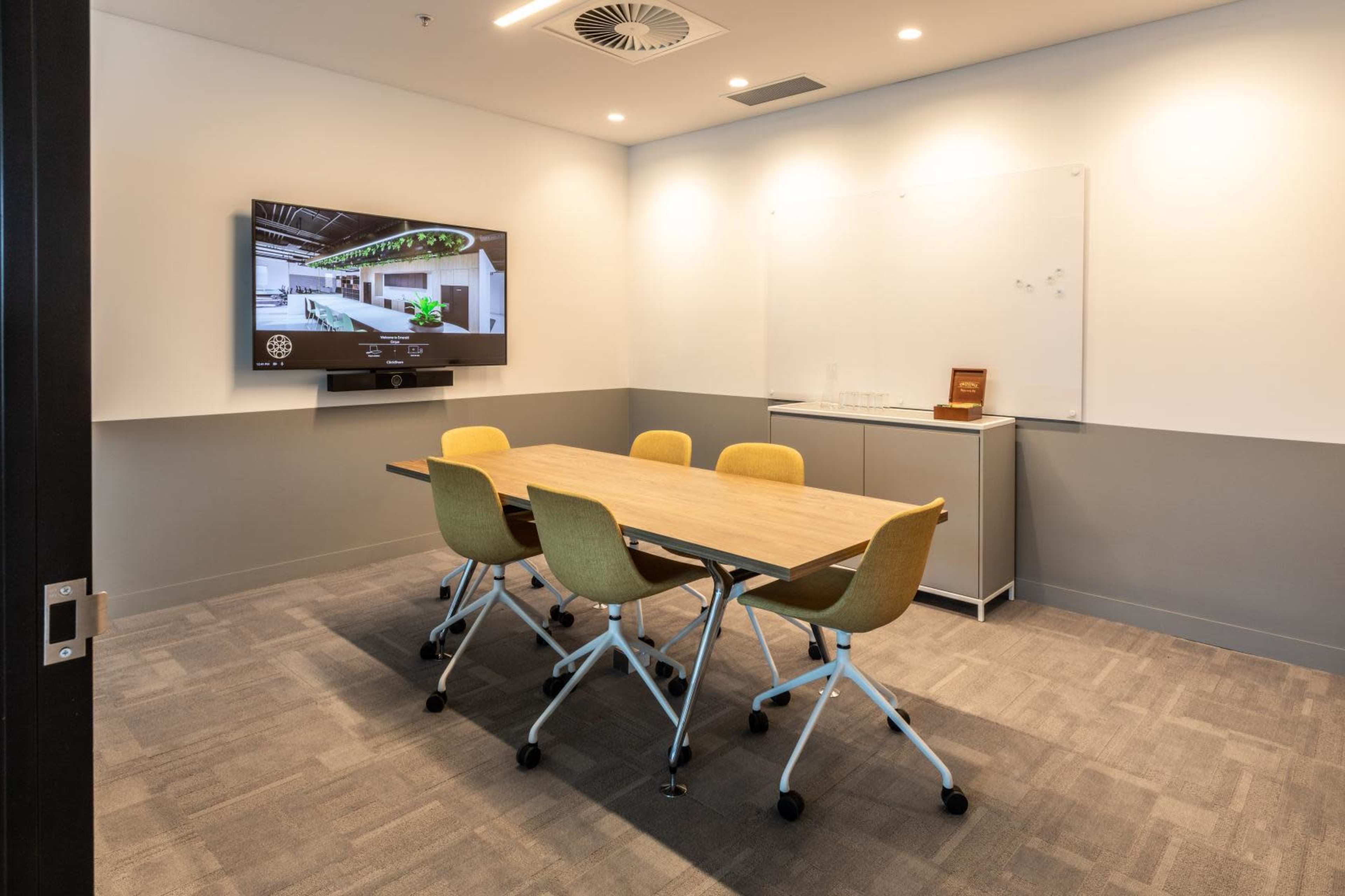 A small conference room features a wooden table surrounded by yellow chairs, a wall-mounted television, and a whiteboard.