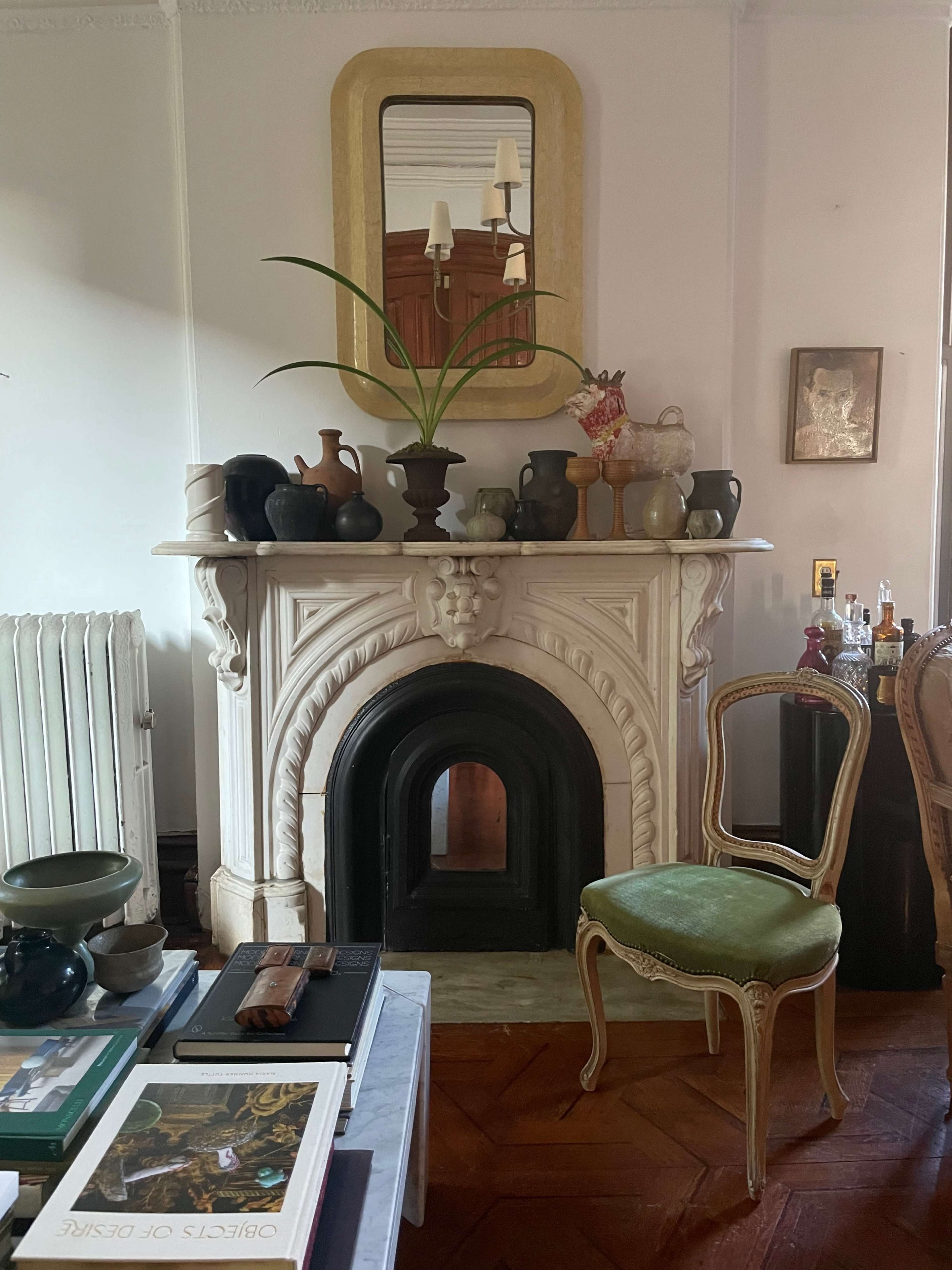 The image shows a fireplace with an ornate mantel, decorated with various pottery pieces and a plant, beside a green chair in a room with wooden flooring.