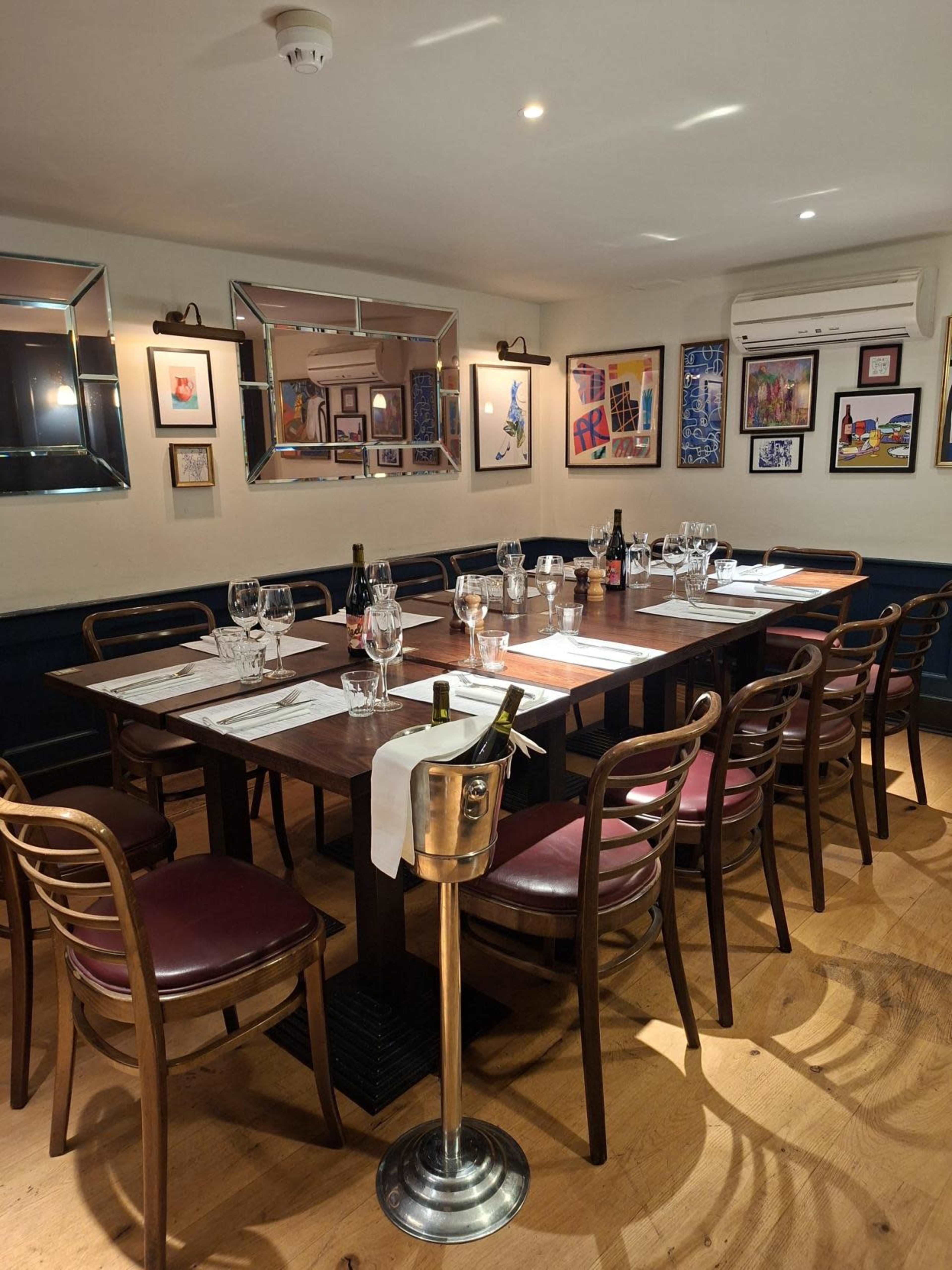 A wooden dining table is set for a meal in a well-decorated restaurant, surrounded by artwork on the walls and featuring chairs with burgundy cushions.