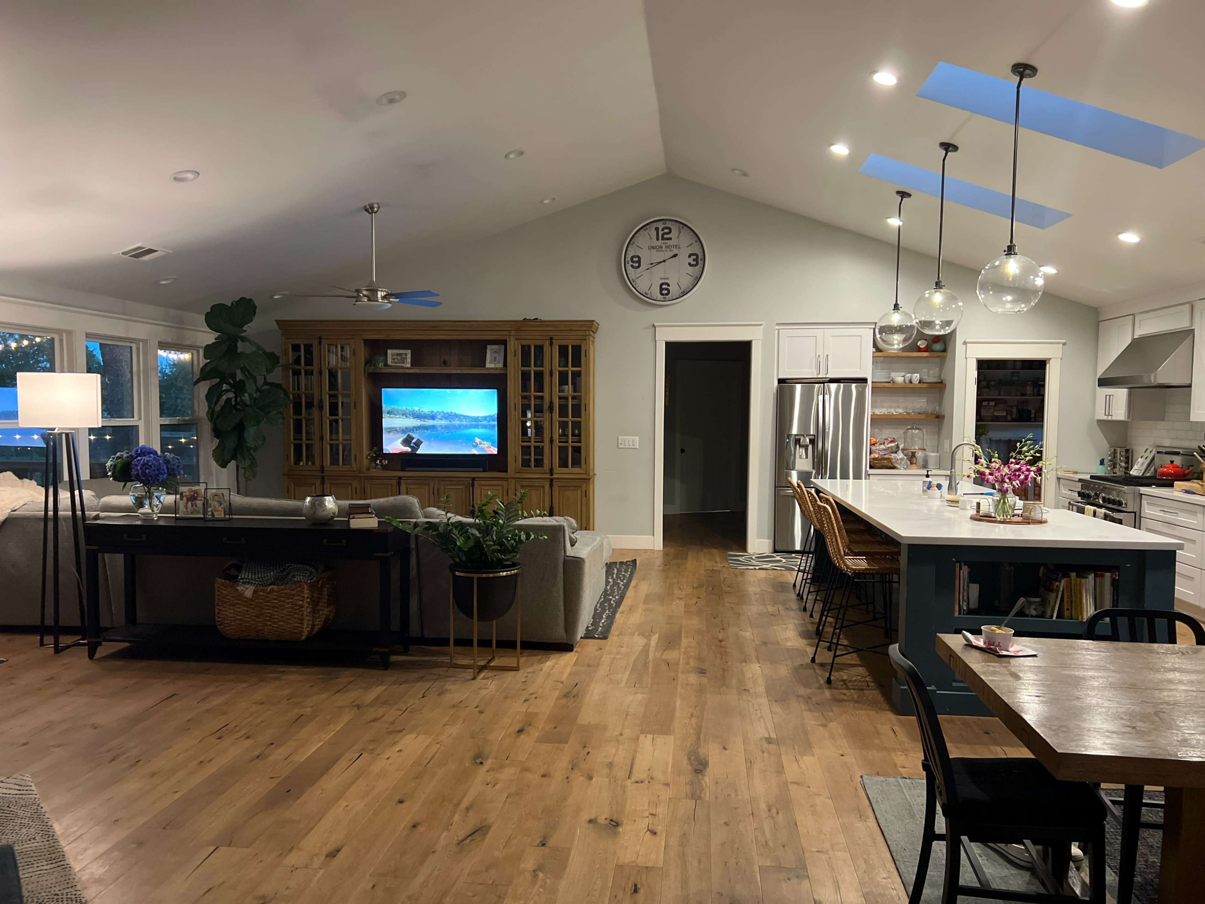 The image shows a modern living room and kitchen area with a large television, a dining table, and an open layout featuring wooden flooring and ample natural light.