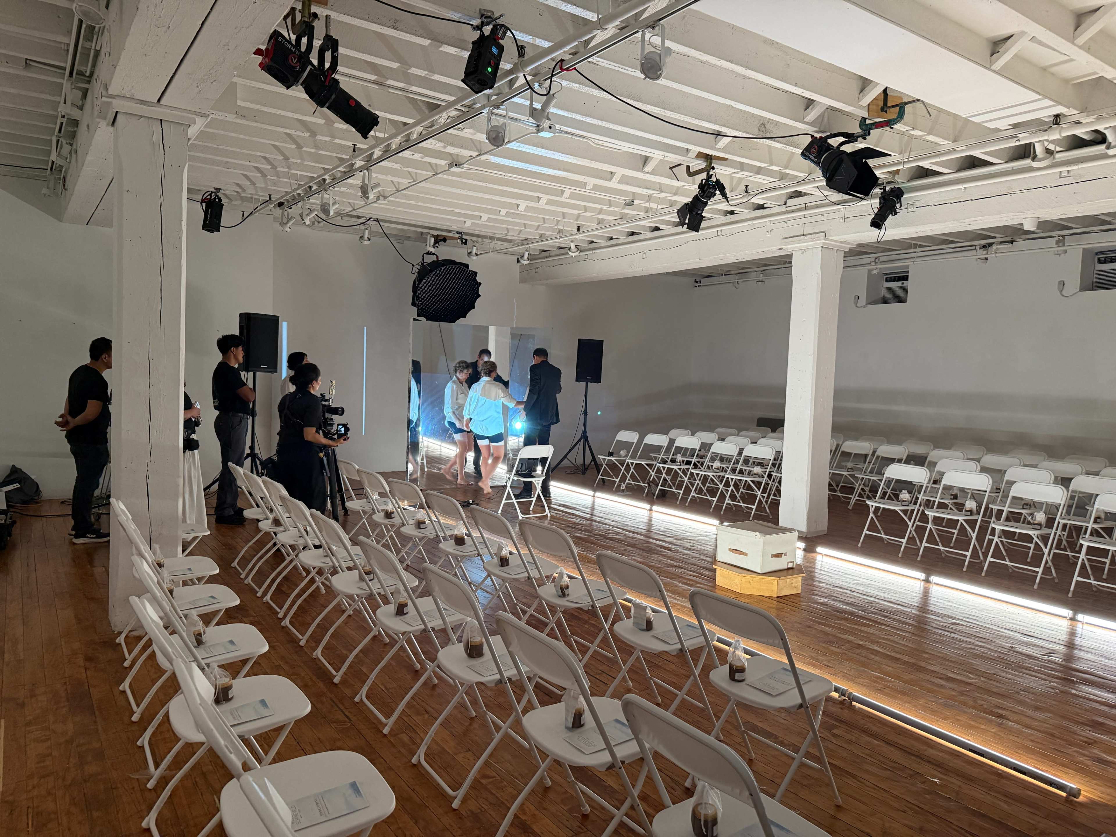 A spacious indoor venue is set up with rows of white chairs facing a stage area, and several people are preparing for an event with equipment and lighting.