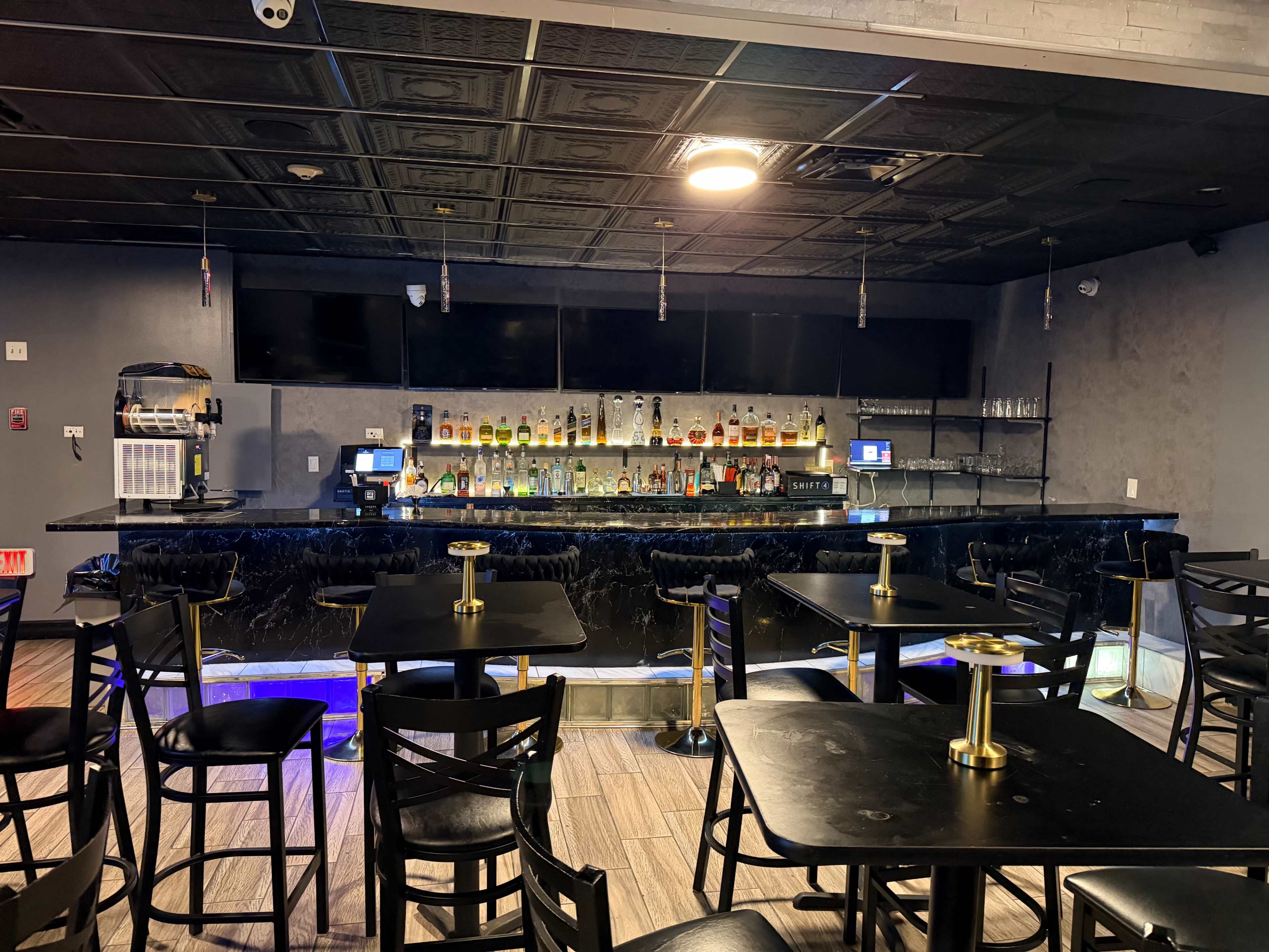 The image shows a dimly lit bar area with a polished black countertop, shelves stocked with liquor bottles, and several tables with black chairs arranged in front.