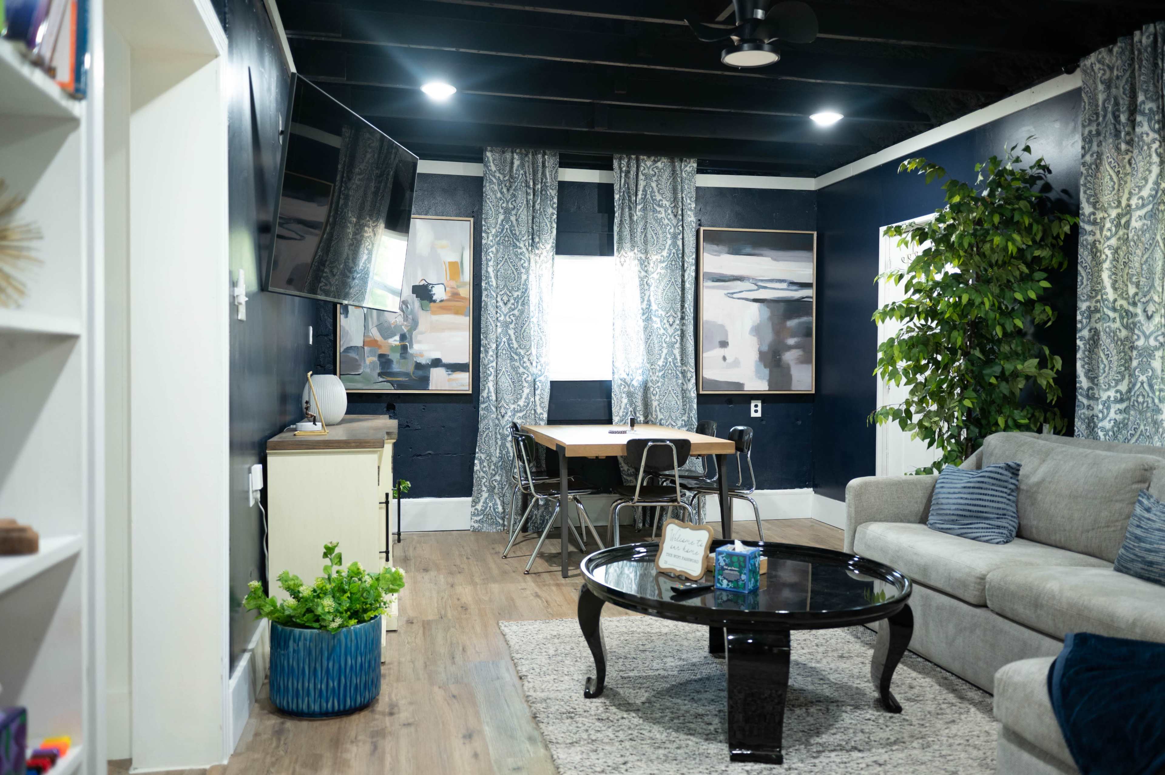 The image shows a living room with a gray sofa, a circular black coffee table, a dining area with a wooden table, and large windows draped with patterned curtains.
