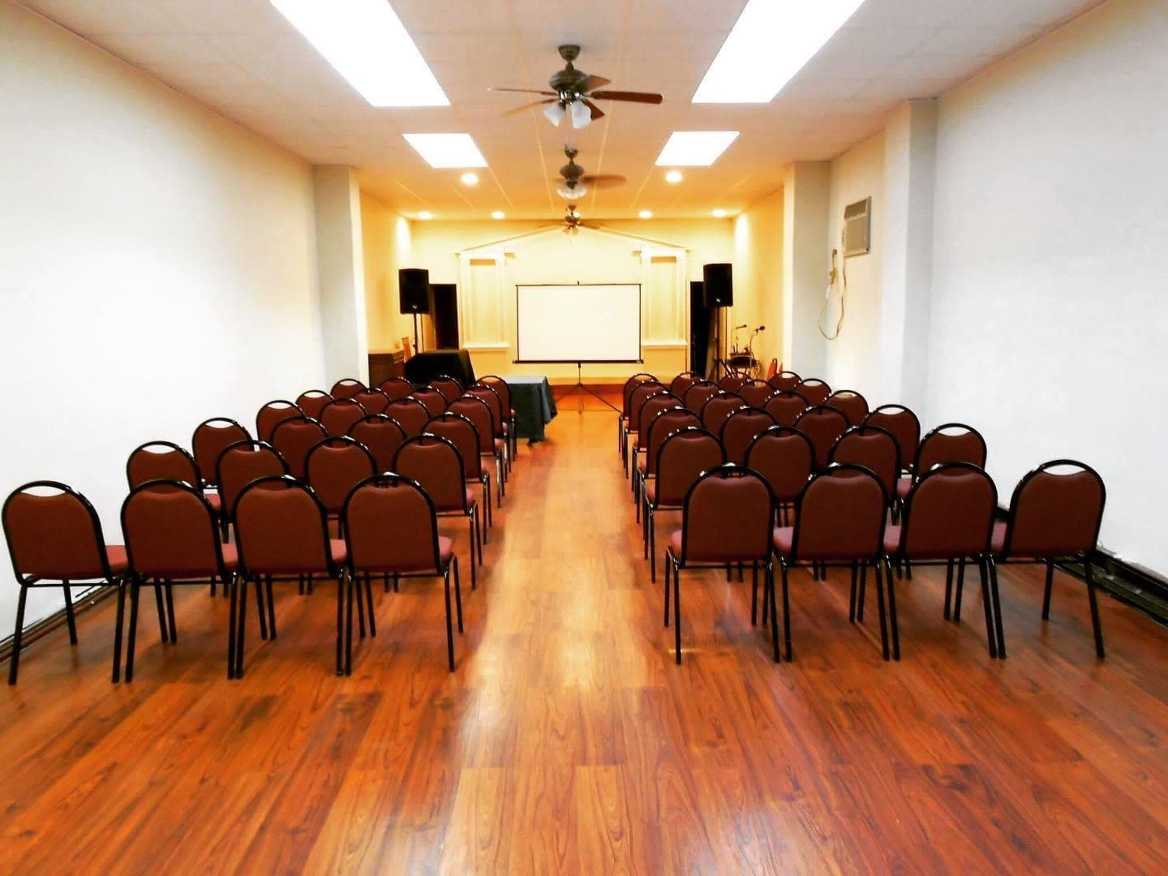 An empty event space arranged with rows of brown chairs facing a blank screen at the front of the room.