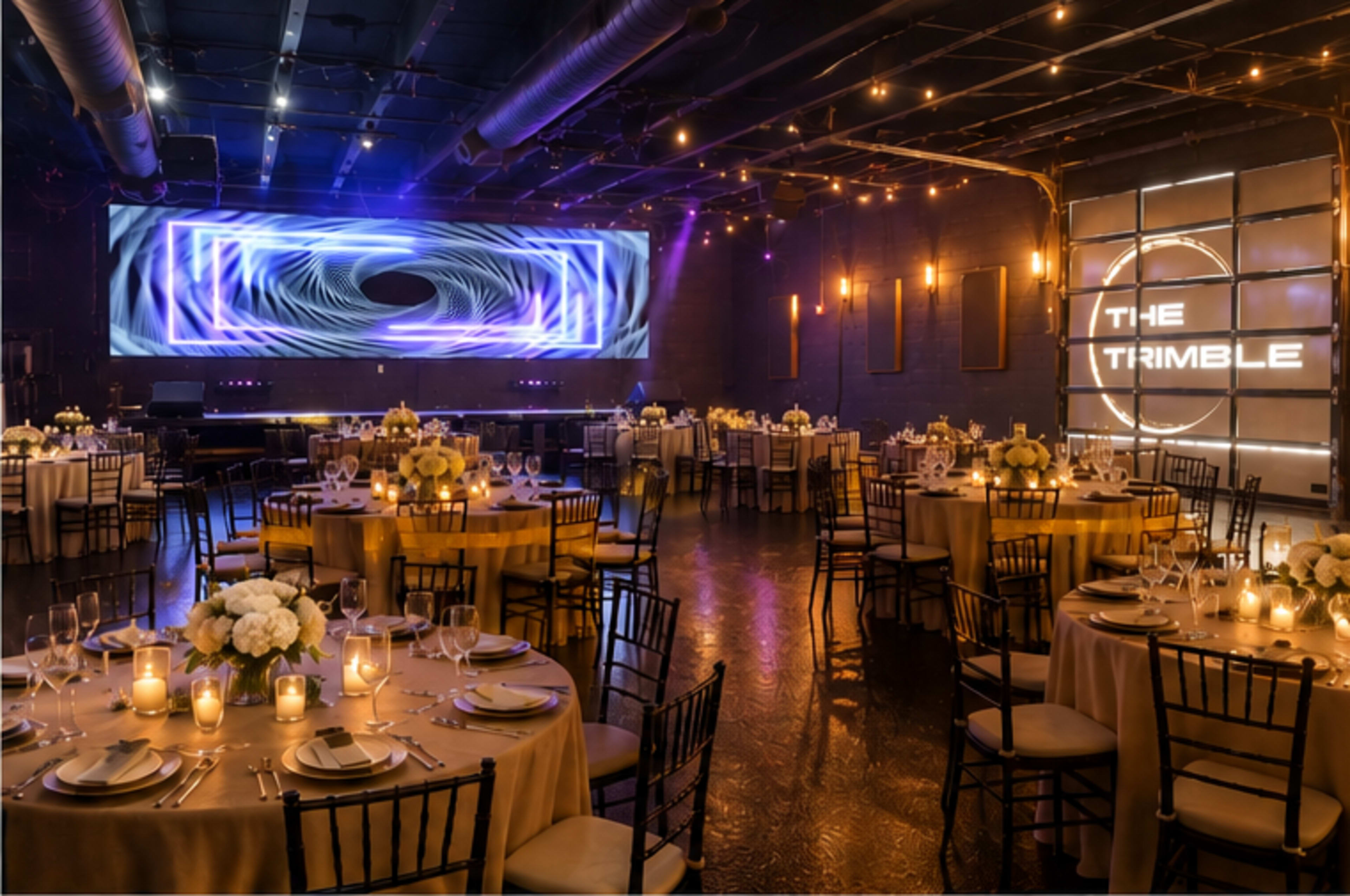 The image shows an elegantly arranged event space with round tables set for a banquet, featuring floral centerpieces and soft lighting, along with a large screen displaying abstract visuals.