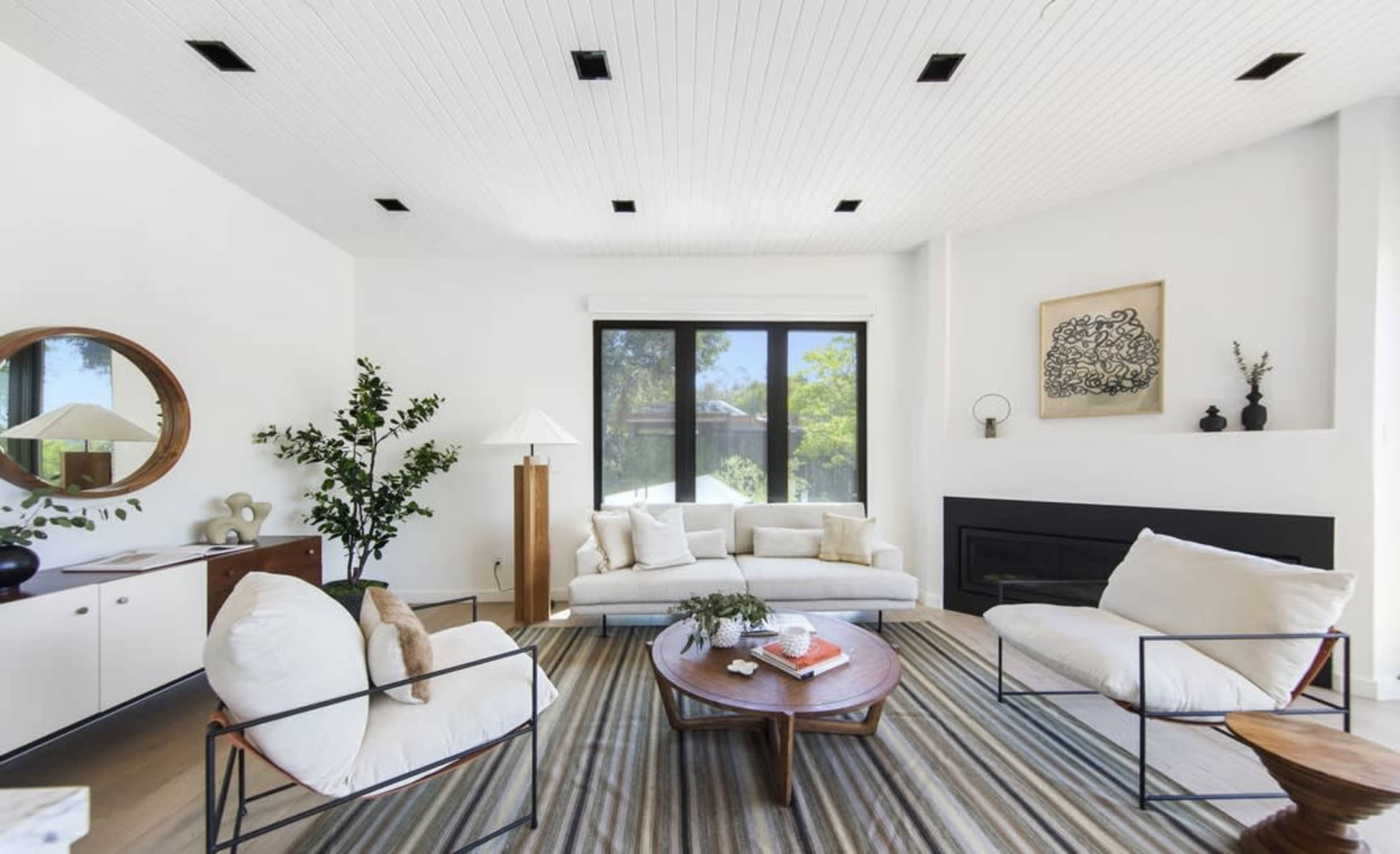 The image shows a modern living room with a white color scheme, featuring a sofa, two armchairs, a round wooden coffee table, and a large window overlooking greenery.