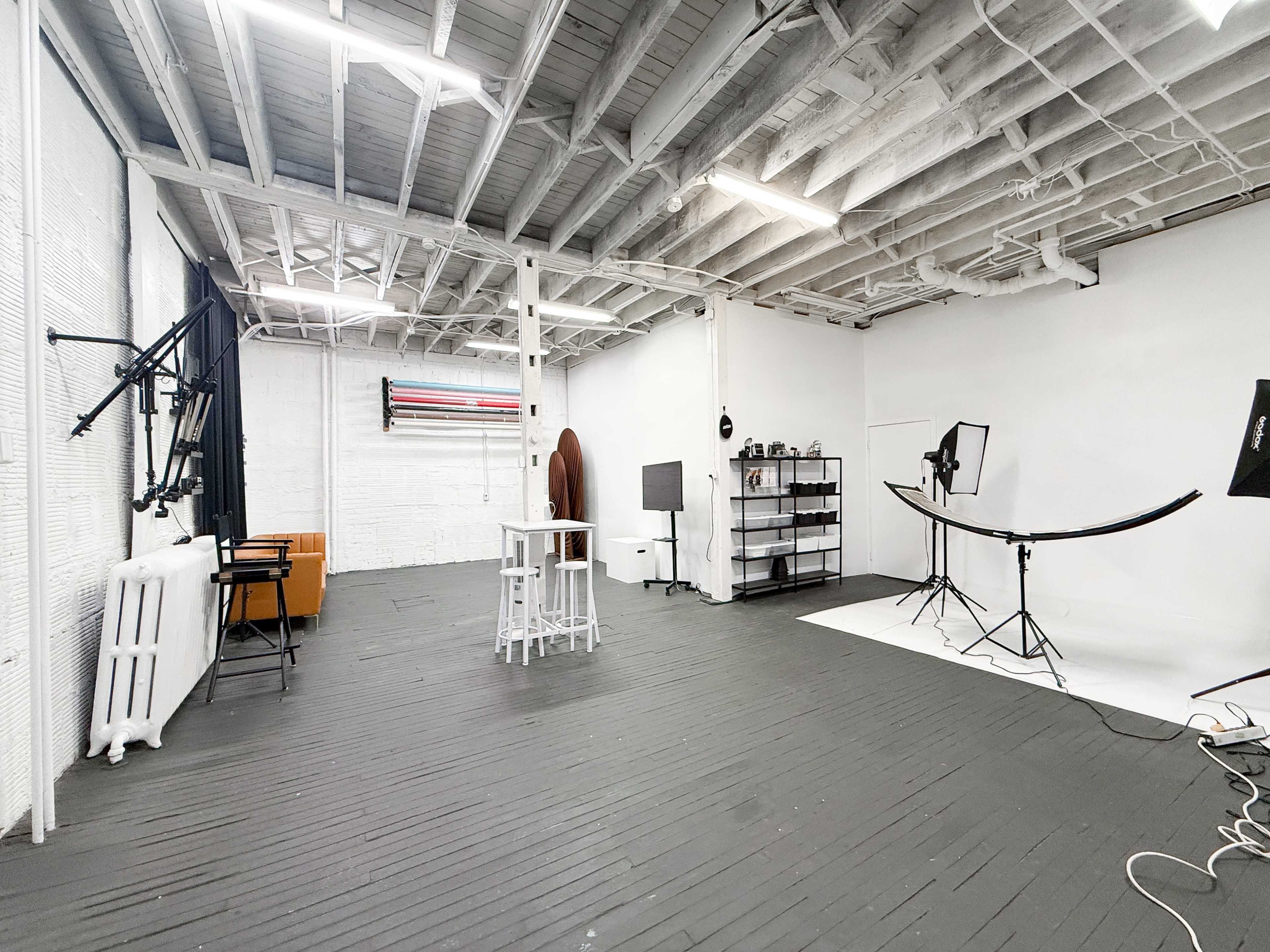 The image shows a spacious photography studio with white walls, a wooden floor, and various equipment including light stands, backdrops, and props arranged neatly.