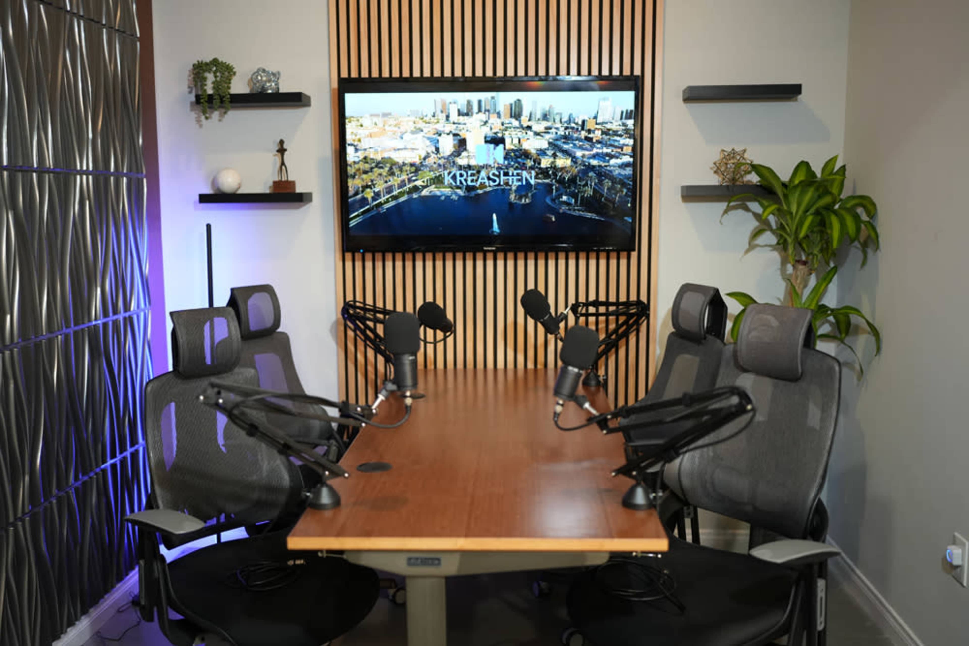 The image shows a modern conference room equipped with a long wooden table, several black chairs with microphones, and a large screen displaying an aerial view of a city.