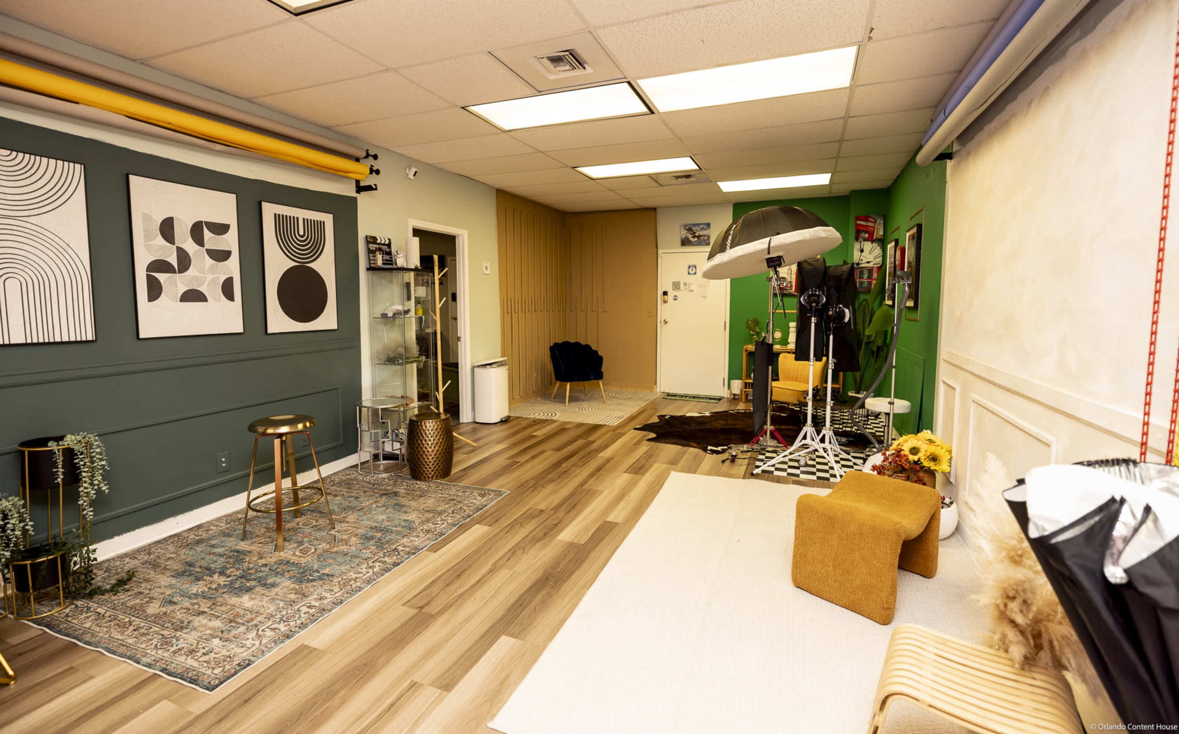 The image shows a spacious studio or creative workspace featuring a combination of green and neutral walls, various art prints, a patterned rug, seating areas, and photography equipment.