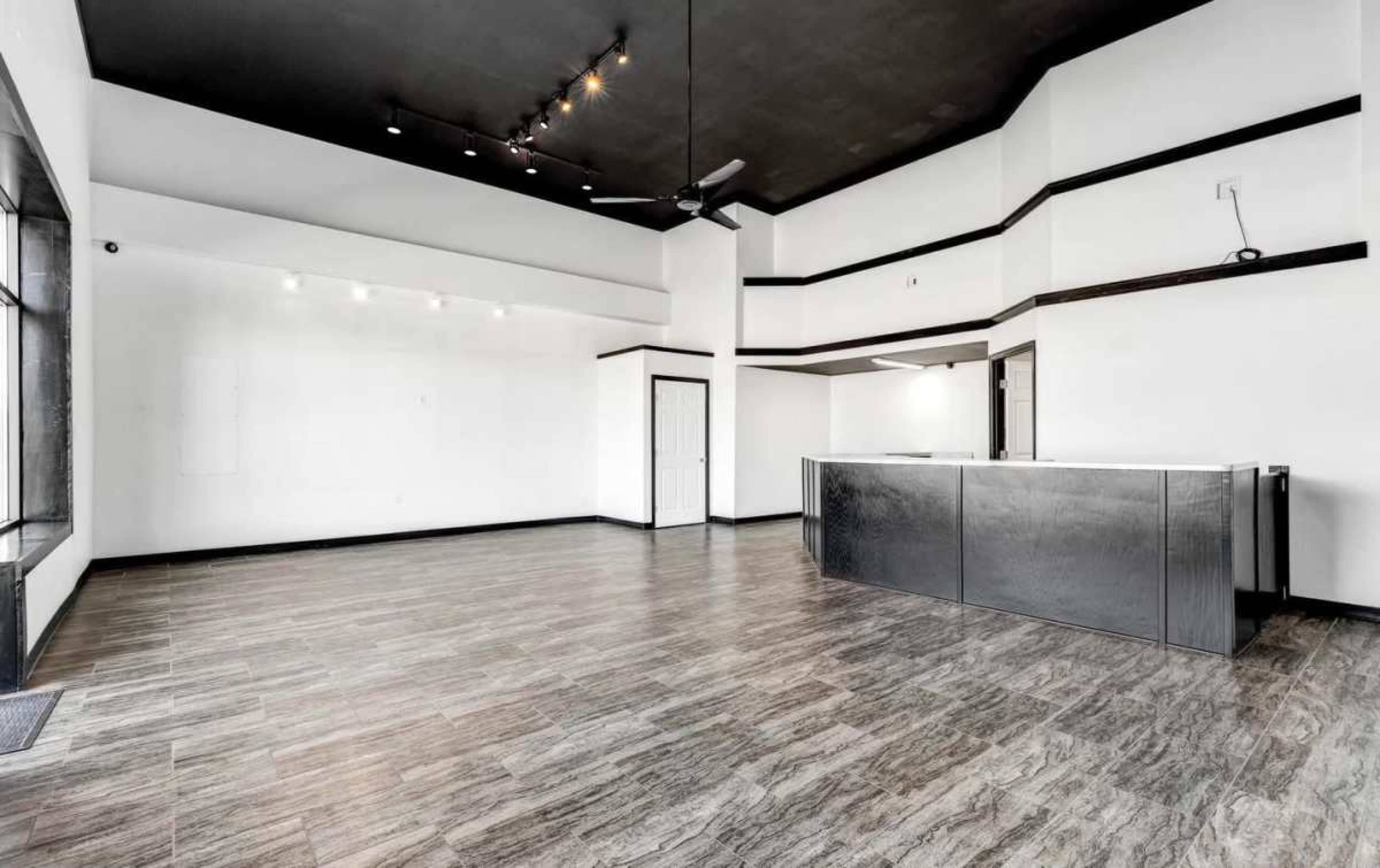 The image shows a spacious, empty room featuring a dark ceiling, a gray reception desk, and wood-like flooring.
