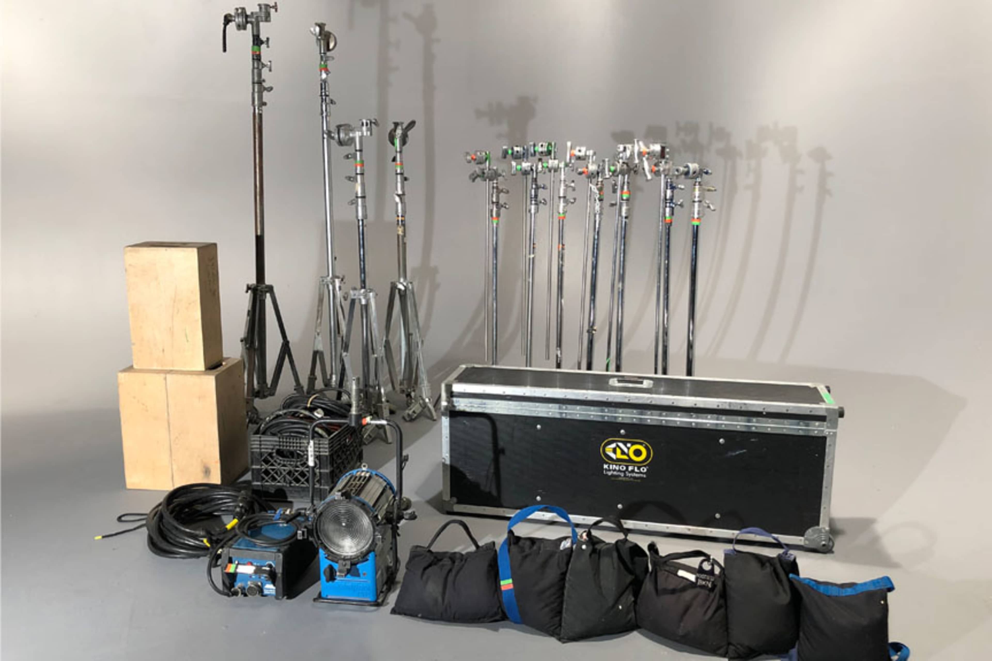 The image shows a collection of film equipment, including lighting stands, crates, cables, a spotlight, and bags arranged on a gray background.