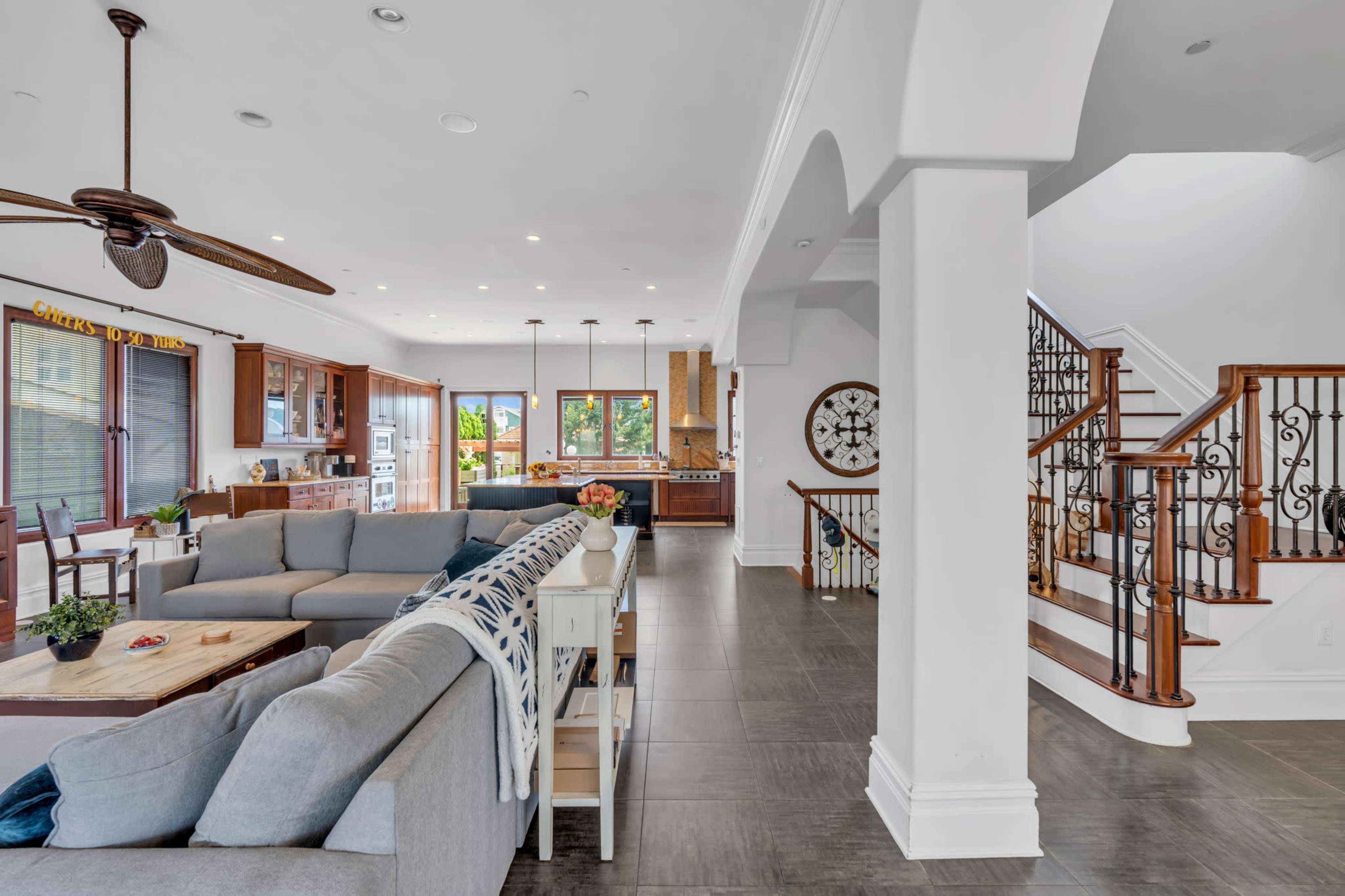 The image shows a spacious living area connected to a kitchen, with a large sectional sofa, wooden dining table, and a staircase leading to an upper level.
