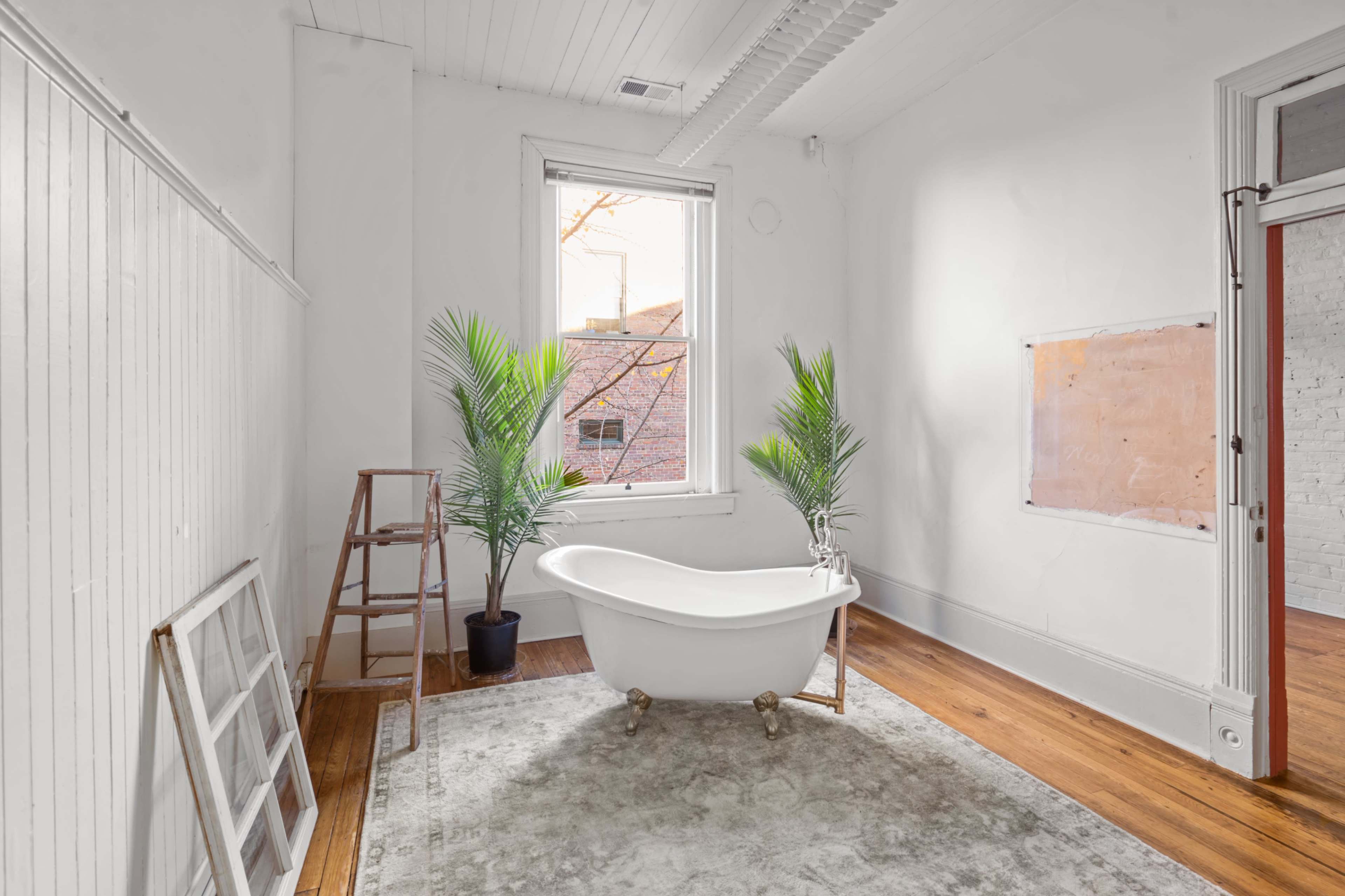 The image shows a bright, minimalist bathroom with a freestanding bathtub, two indoor plants, and wooden flooring.