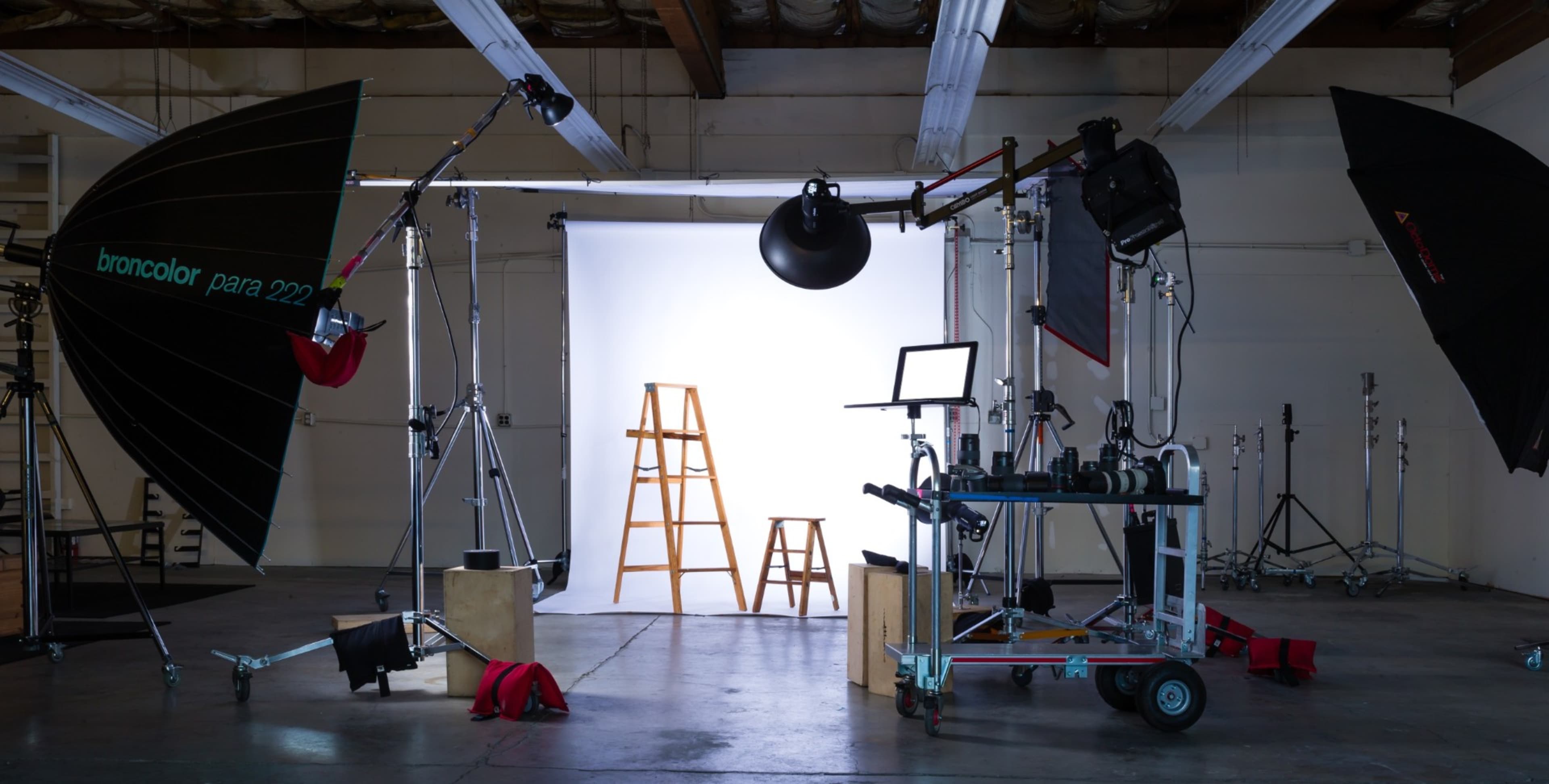 A photography studio setup with two ladders, multiple light sources, and various equipment arranged around a backdrop.