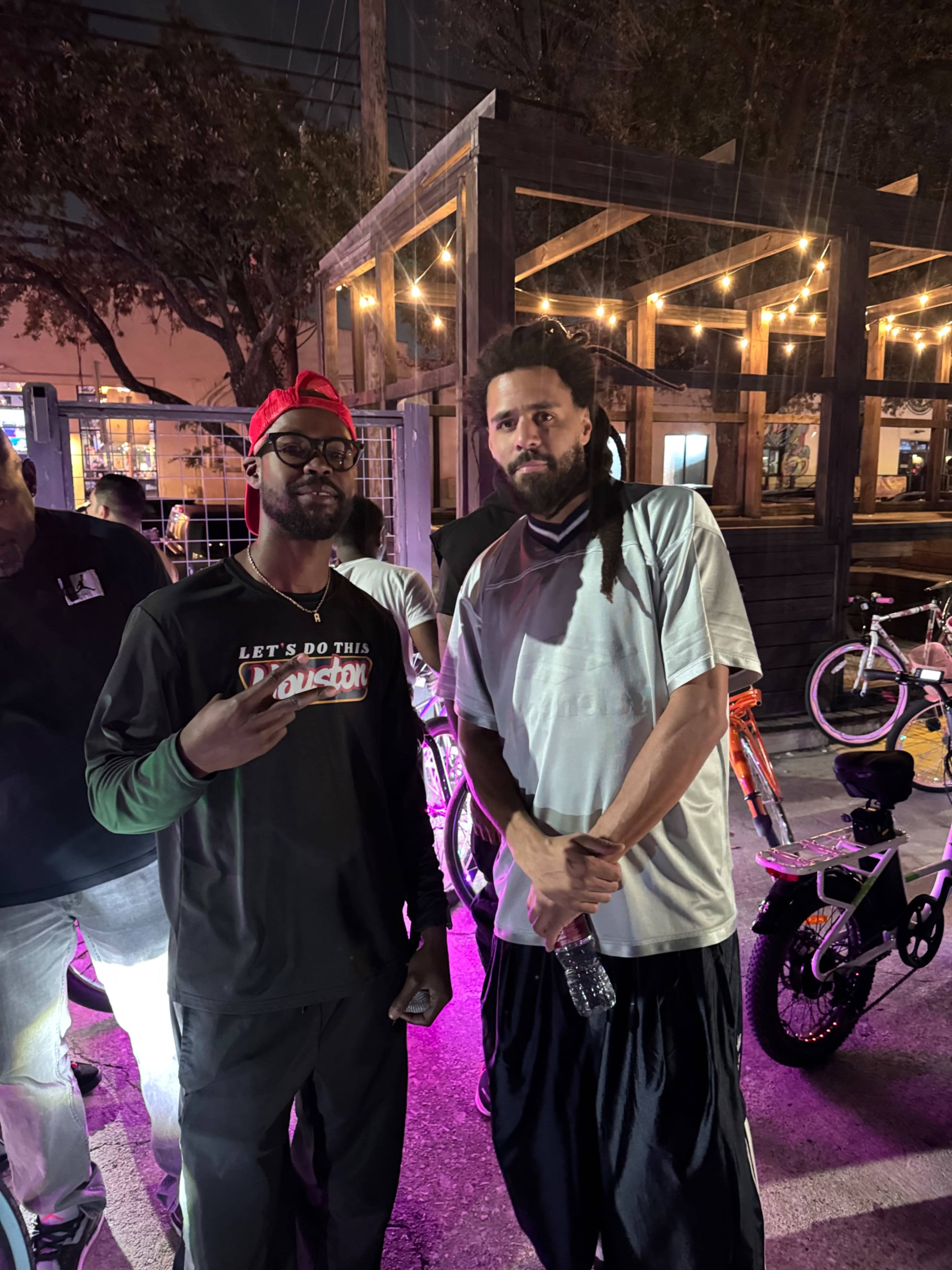 A man wearing glasses and a black shirt poses with another man in a white shirt, both standing in a dimly lit outdoor setting with bicycles nearby.